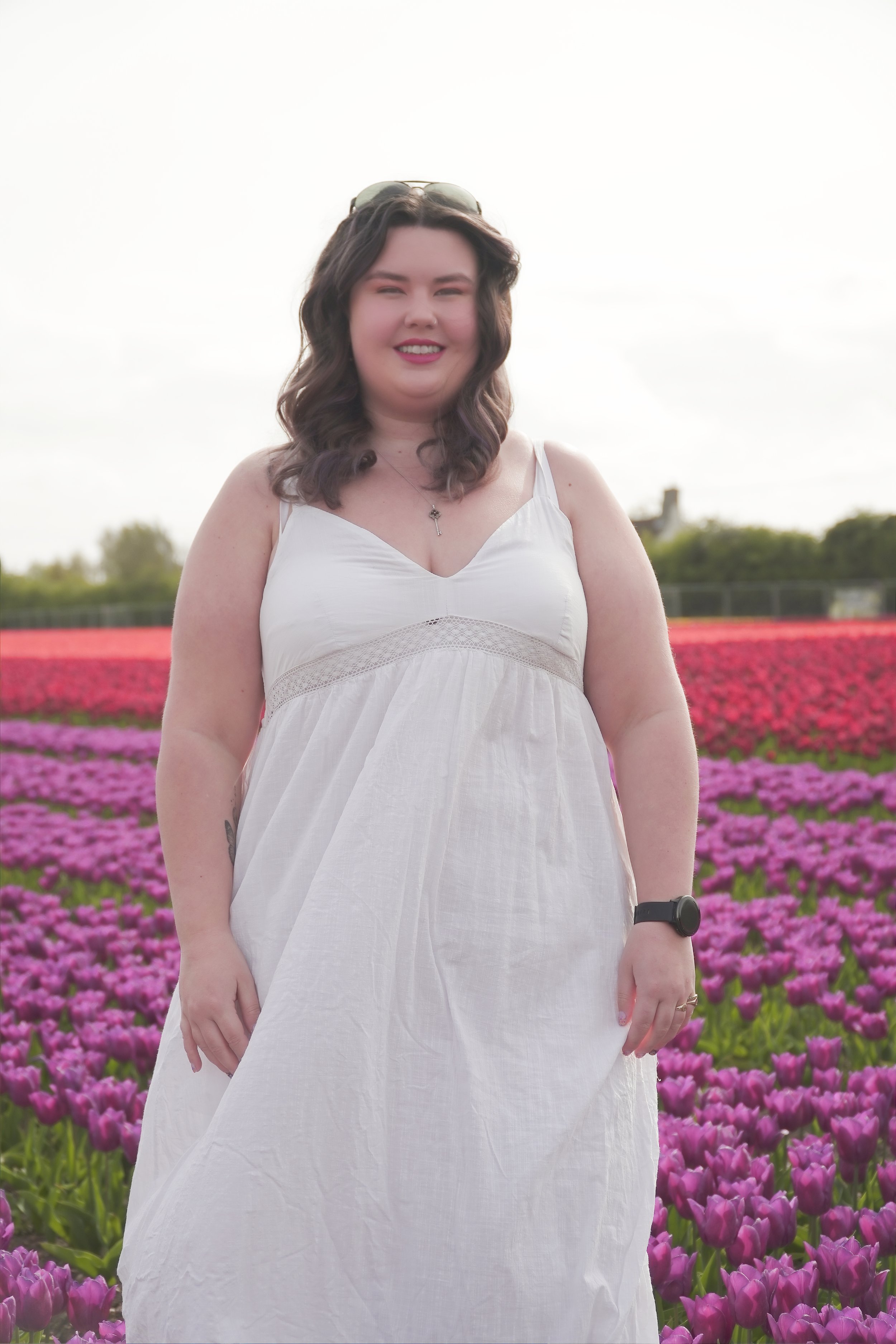 A woman standing in a field of blooming pink and purple tulips on a cloudy day, wearing a white sundress, sunglasses on her head, and a watch on her left wrist, smiling at the camera.