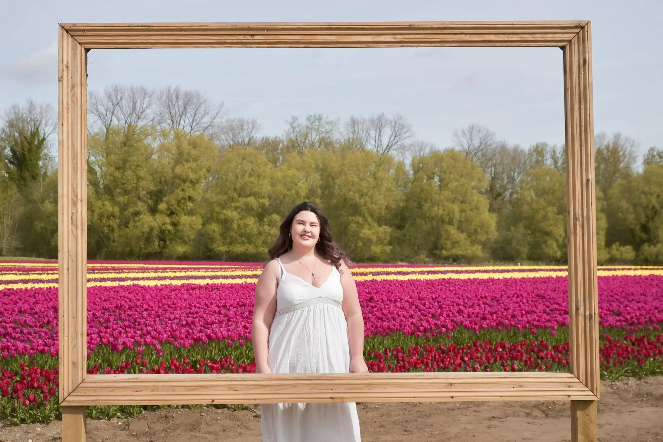 A woman in a white dress standing in front of a tulip field, framed by a large wooden rectangular frame, with trees and a partly cloudy sky in the background.