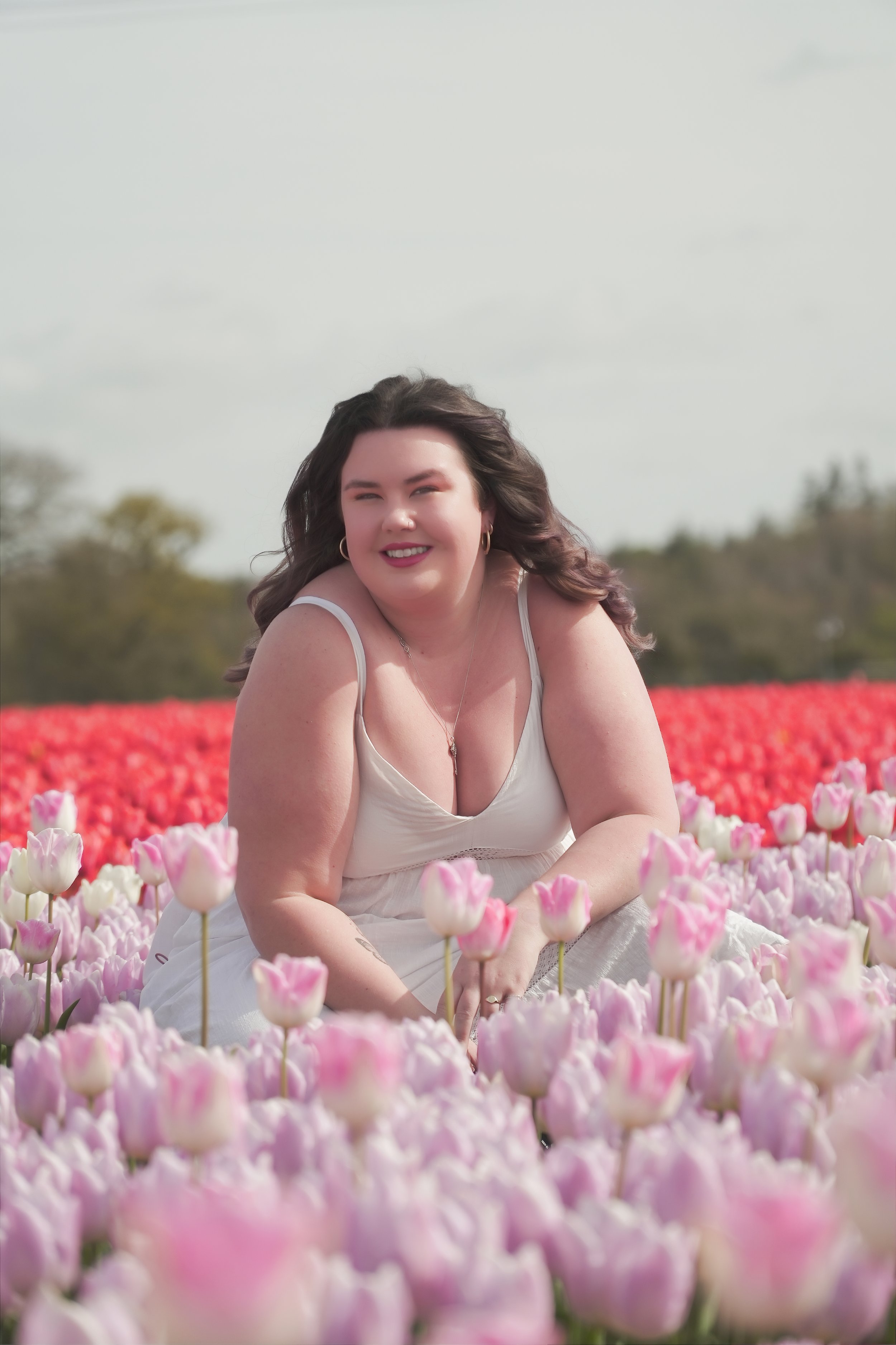 A woman smiling and squatting in a field of pink and white tulips during daytime.