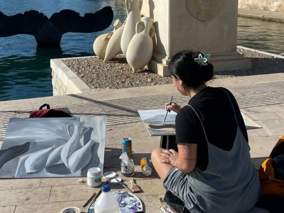 Una mujer pintando en la calle junto a una pintura de objetos blancos. Tiene cabello oscuro atado con una cinta y está sentada en el suelo con materiales de pintura a su alrededor. Detrás hay una fuente con esculturas de vasijas y una estructura de piedra junto al agua.