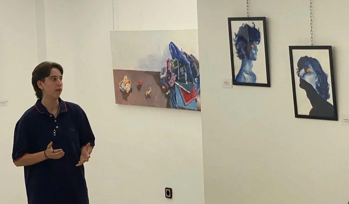Una mujer en una galería de arte observando varias pinturas en las paredes, que incluyen retratos en estilo artístico y una escena con objetos sobre una mesa.