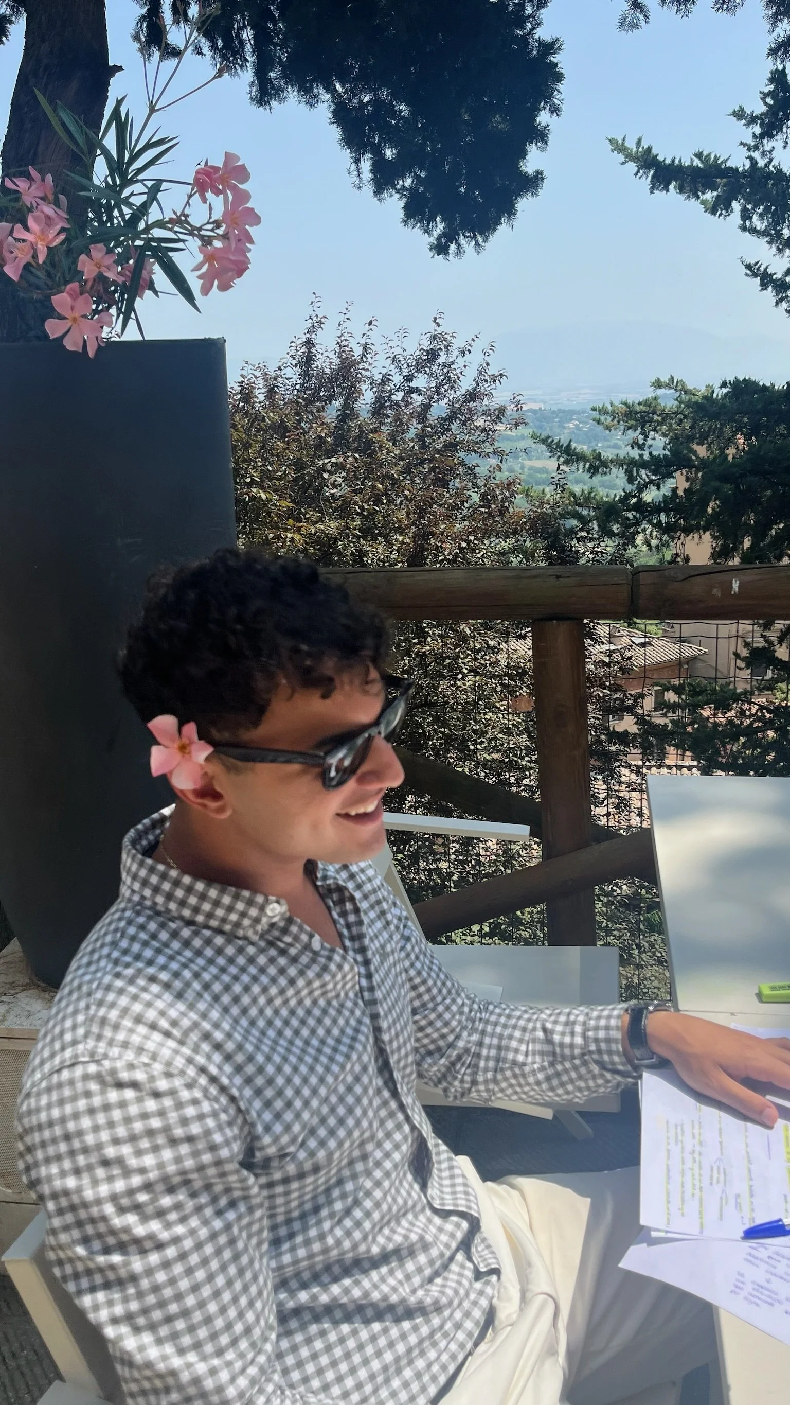 Un hombre con gafas y camisa de cuadros se encuentra sonriendo, sentado en una terraza, trabajando con papeles y una laptop, con un paisaje de árboles y ciudad al fondo.