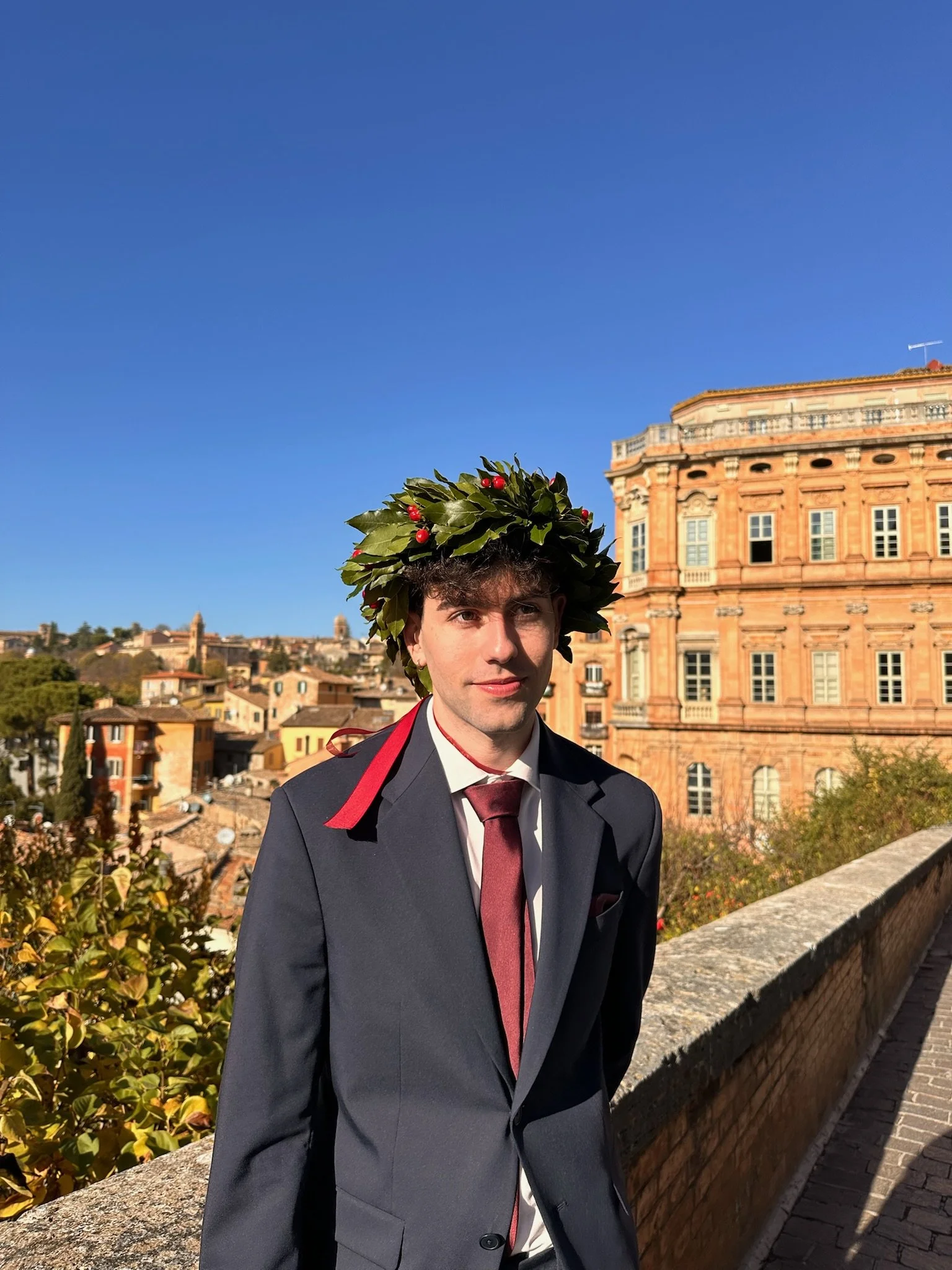 Joven con toga y laurel en la cabeza en un lugar con edificio histórico y cielo azul.