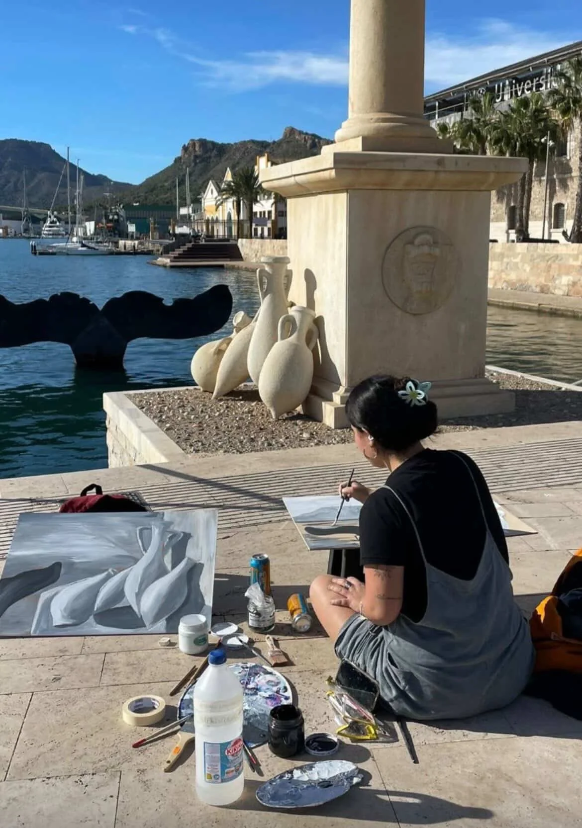 Una artista pintando en el malecón frente a un cuerpo de agua y montañas en el fondo. La artista tiene cabello oscuro y lleva puesta una camiseta negra y pantalones cortos grises. En su alrededor hay pinturas, pinceles, latas, y otros materiales de arte sobre la superficie de suelo de piedra. Detrás, hay estatuas de cerámicas y una estructura con columnas y el logotipo de una universidad.