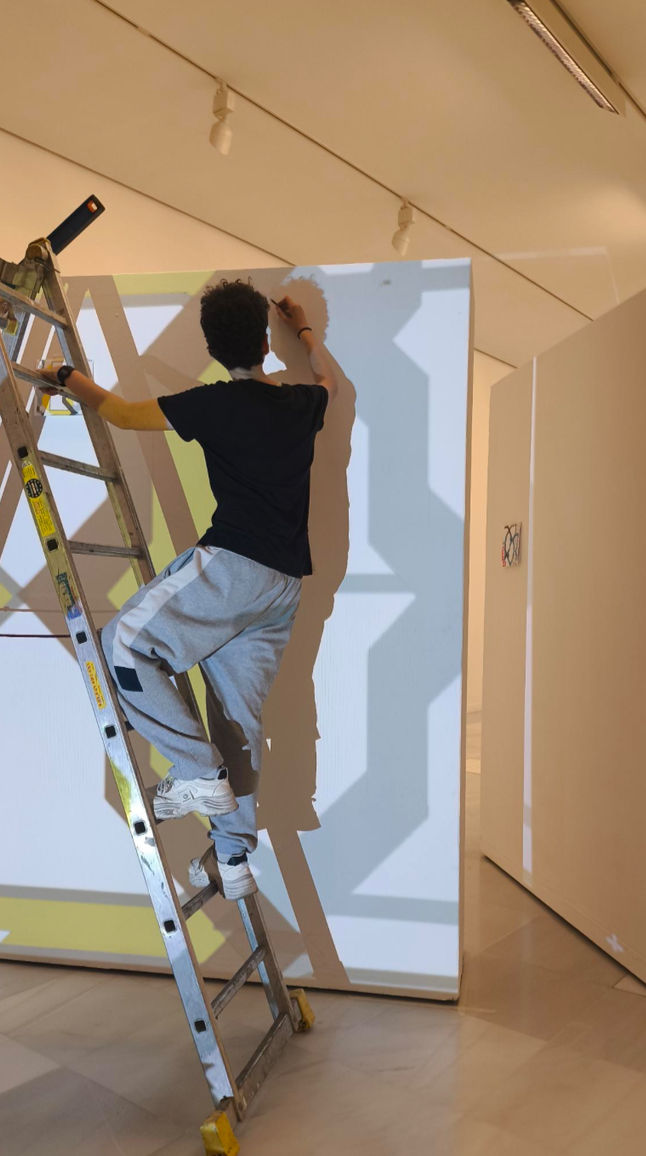 Una persona de espaldas pintando una pared con un pincel, usando una escalera y con sombras en la pared, en un espacio interior con iluminación artificial.