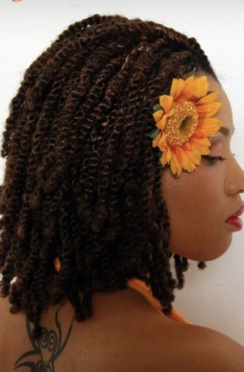 A woman with brown textured locs wearing a sunflower hair clip on the side of her head, with a tattoo on her shoulder.