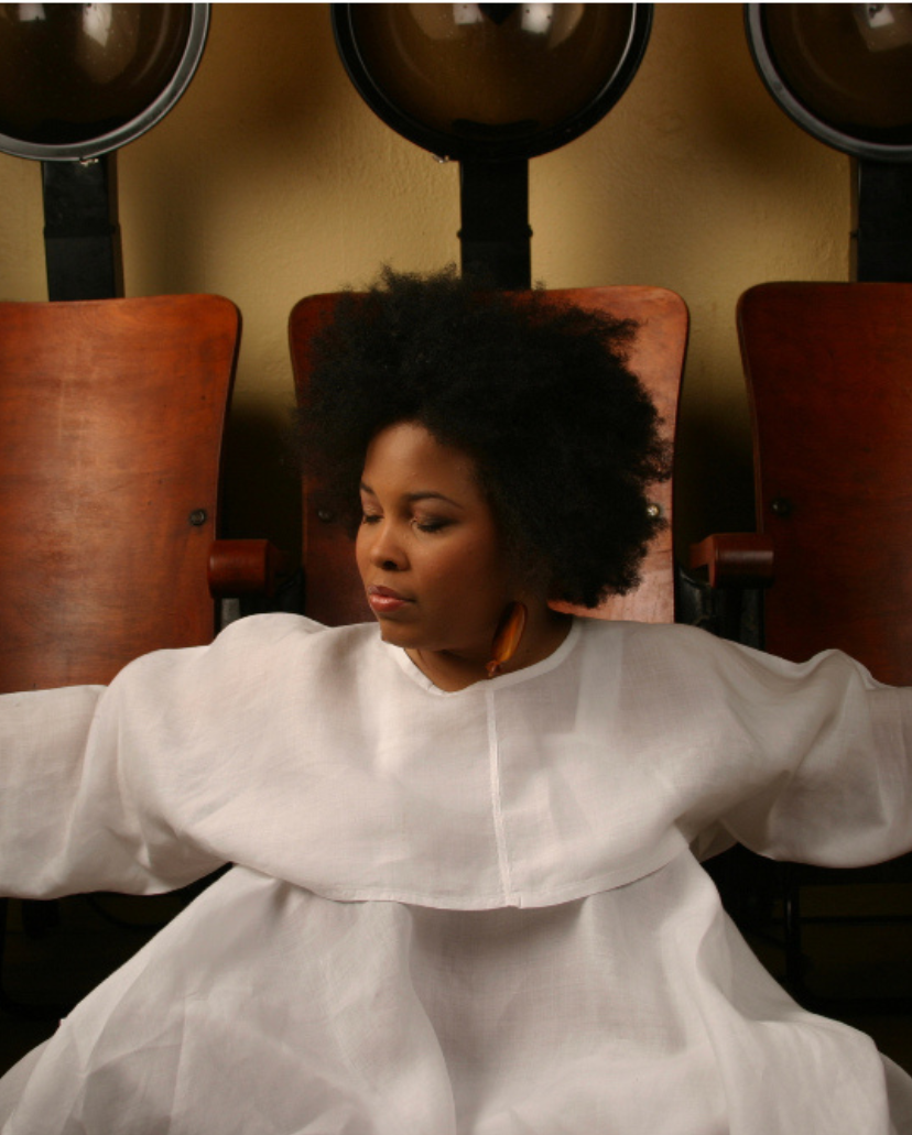Madam Tonya with natural curly hair sitting with closed eyes in a white gown, in a room with vintage wooden chairs and large black hair dryers in the background.