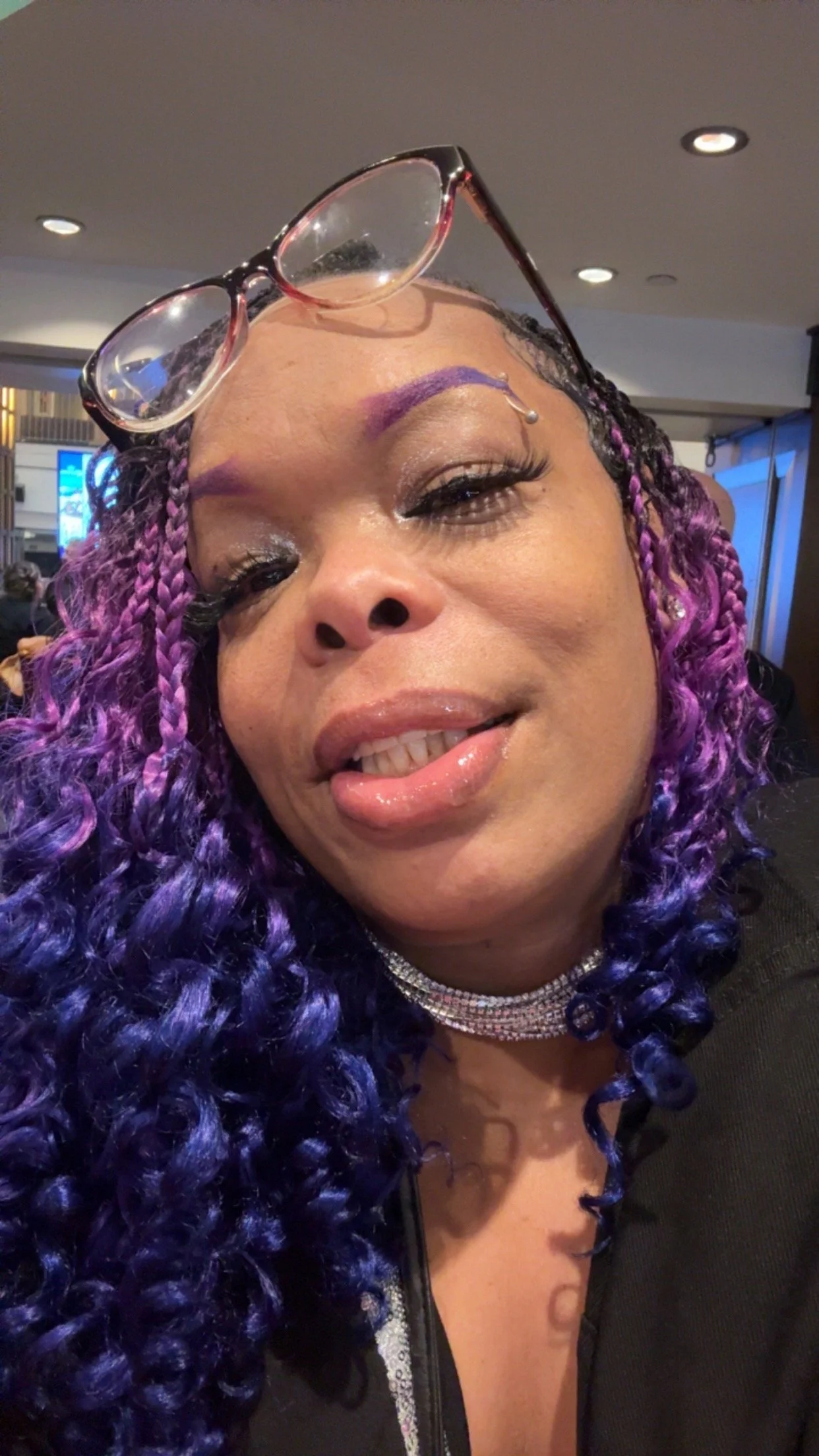 Close-up of a woman with purple and black curly hair, wearing sunglasses on her head, a sparkling choker, and makeup with shiny lipstick and eyeliner.
