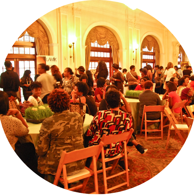People gathered in a grand, elegant room with high ceilings, large arched windows, and gold curtains, sitting at round tables with centerpieces, some standing, others seated, engaging in conversations.
