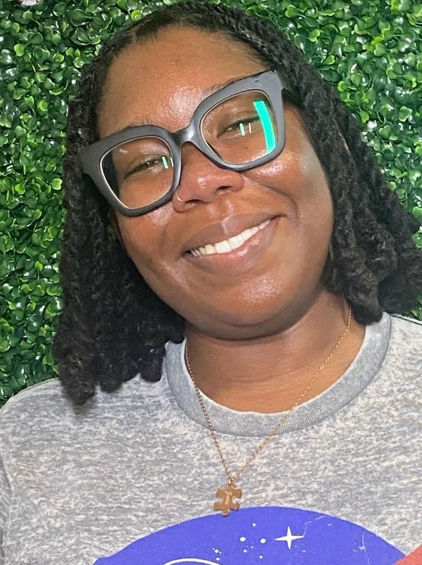 A woman smiling, wearing large glasses, a gray t-shirt, a gold necklace with a puzzle piece pendant, and standing in front of a green leafy background.