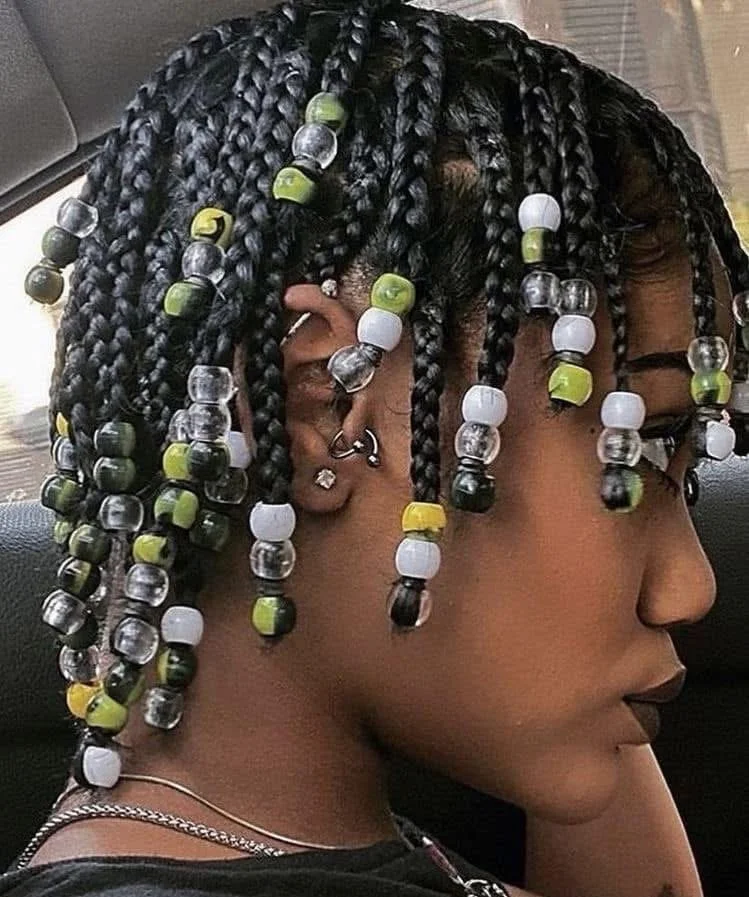 Side profile of a person with styled micro braids decorated with multicolored beads, multiple earrings, and a necklace.