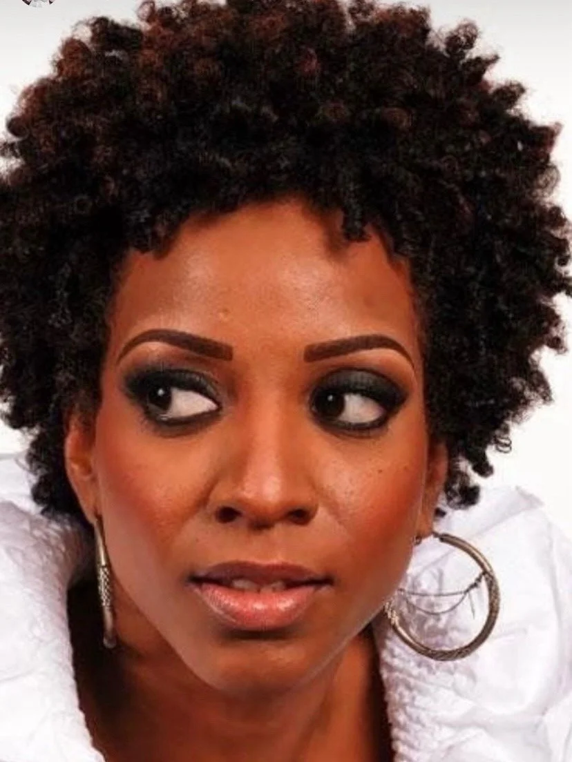 A woman with short, curly hair, wearing makeup, hoop earrings, and a white garment.
