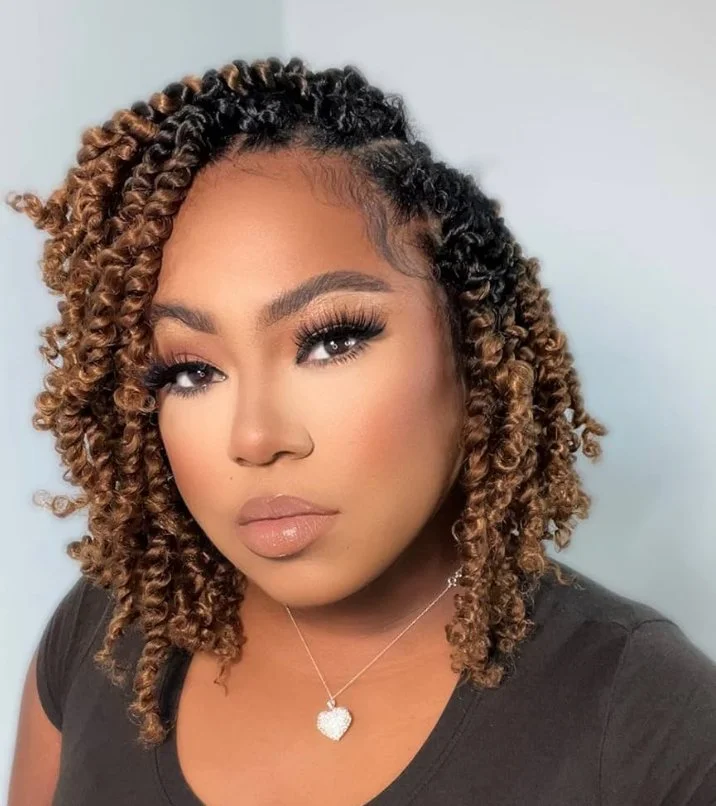 A woman with curly, brown and black streaked hair, wearing makeup including long lashes and nude lipstick, in a black shirt, with a heart-shaped pendant necklace, looking at the camera.