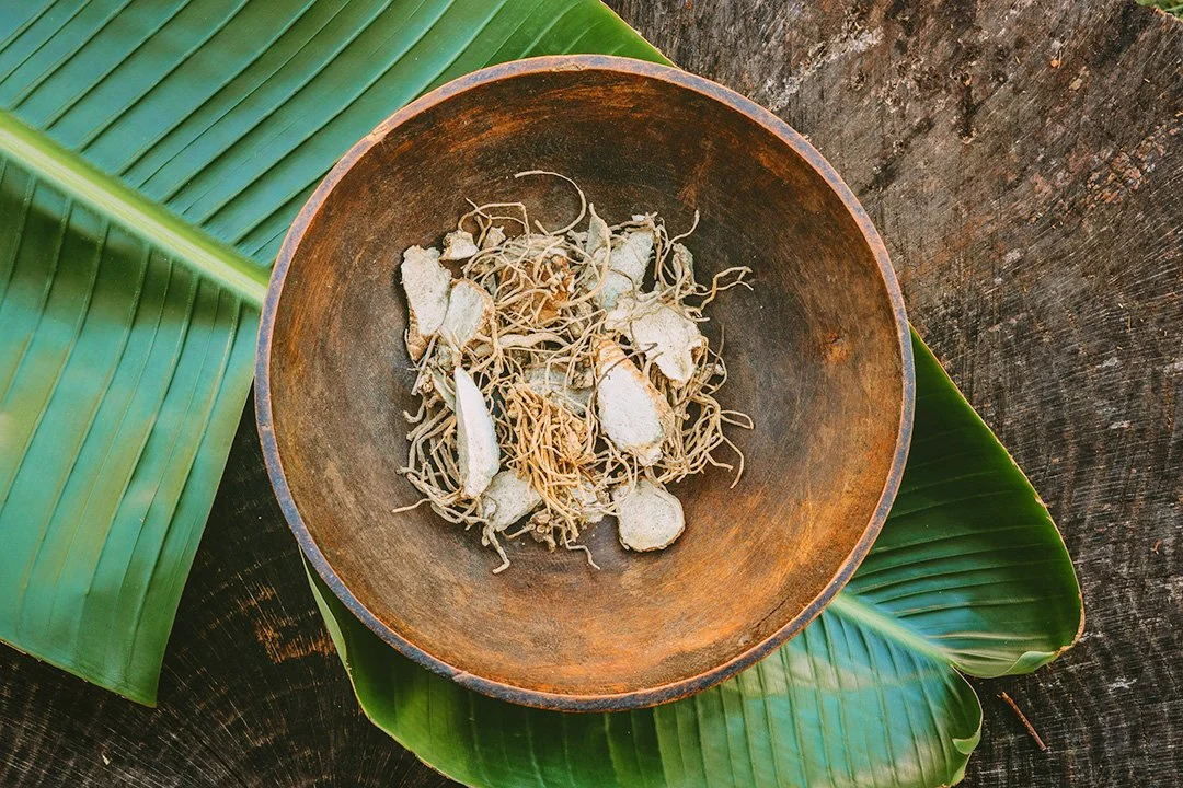 A round wooden bowl containing dried roots and ginger slices, placed on two large green tropical leaves on a dark wooden surface.