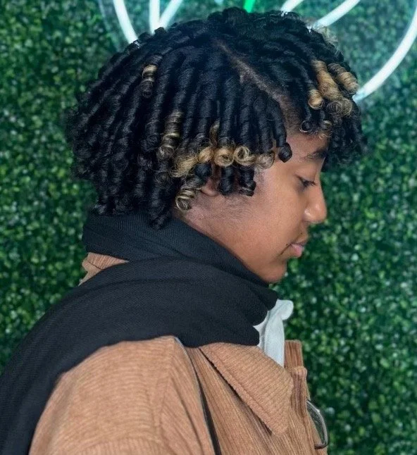 Profile of a person with curly, black and blonde-tipped hair, wearing a brown jacket and black hoodie, against a green leafy background.