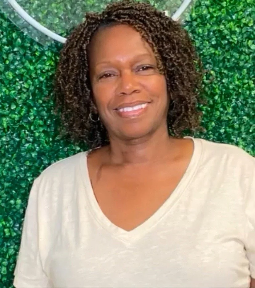A woman with shoulder-length curly hair smiling against a green leafy background.