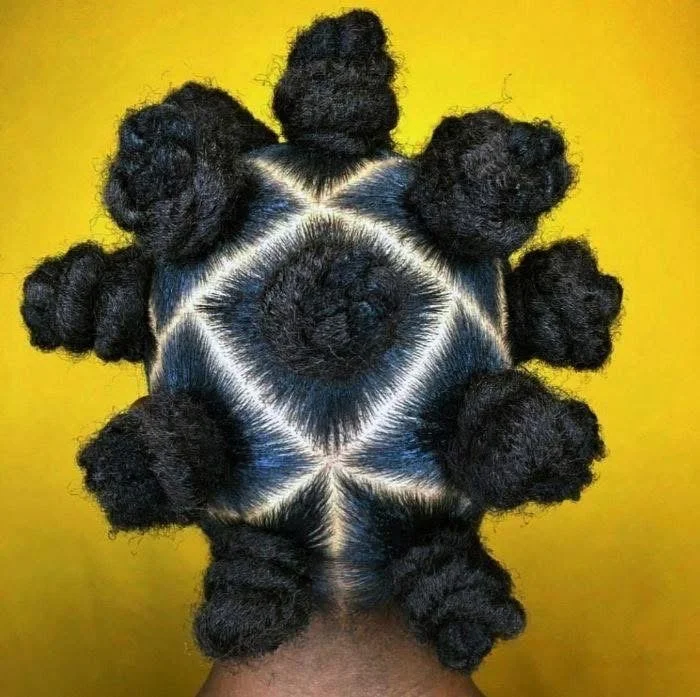 A person's hair styled in symmetrical, circular bantu knots with a triangular pattern of white dye on the scalp, set against a yellow background.
