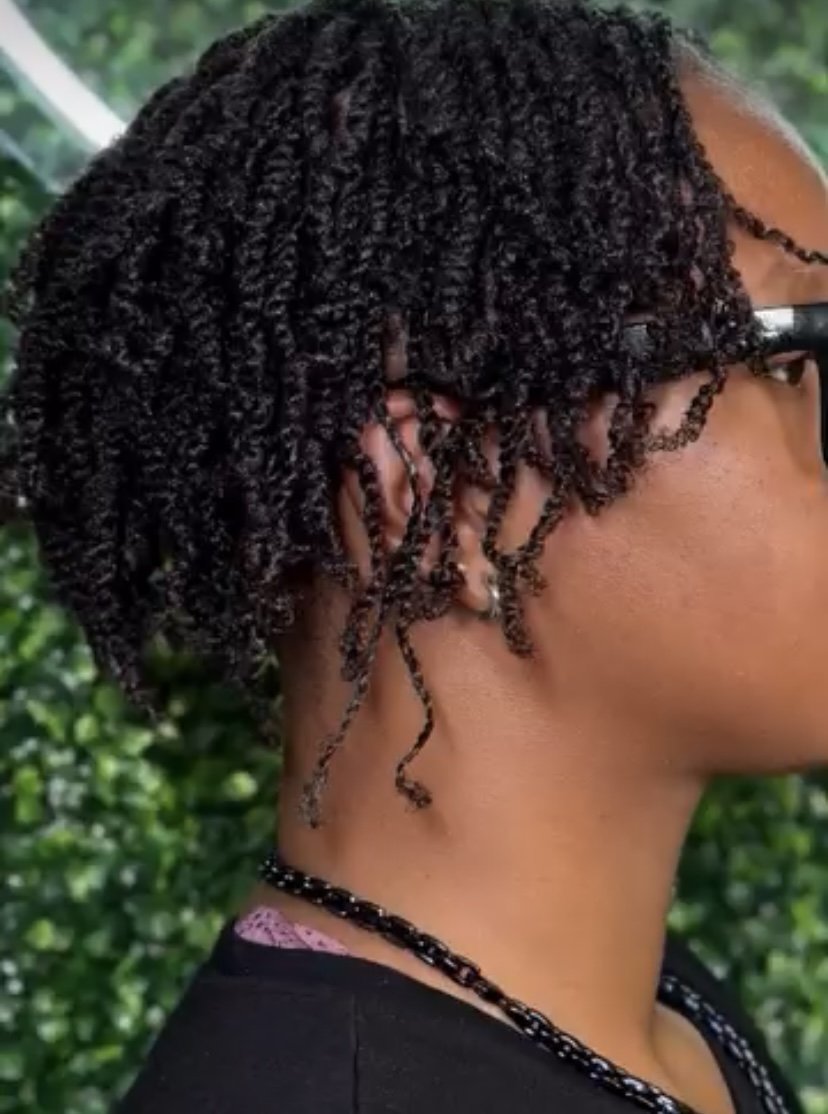 Close-up of a person's head showing short, curly, twisted black hair, with some curls loosely hanging, wearing glasses, a pearl earring, a black beaded necklace, and a dark shirt, with green foliage in the background.