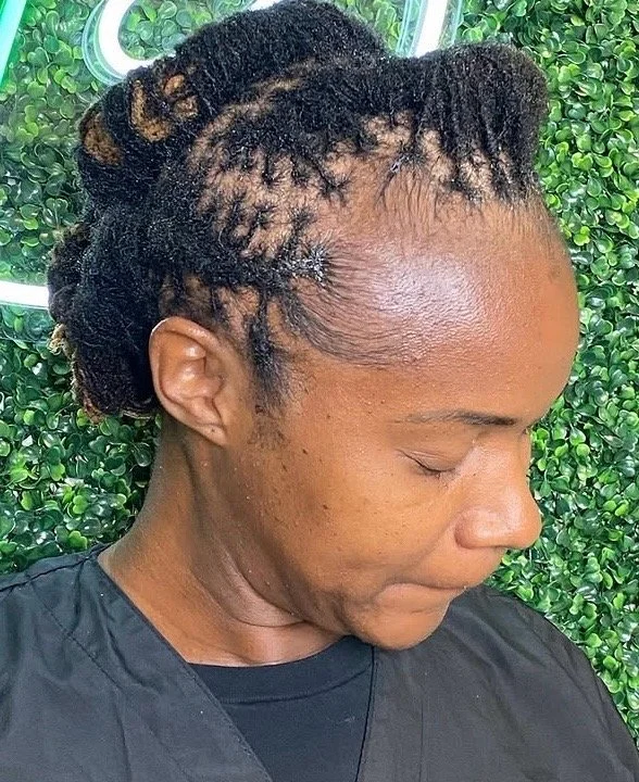 Side profile of a woman with styled dreadlocks, standing in front of a green leafy background.