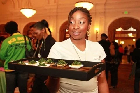 A smiling woman in a white shirt holding a black tray with small dishes of food, at an indoor event with people in the background.