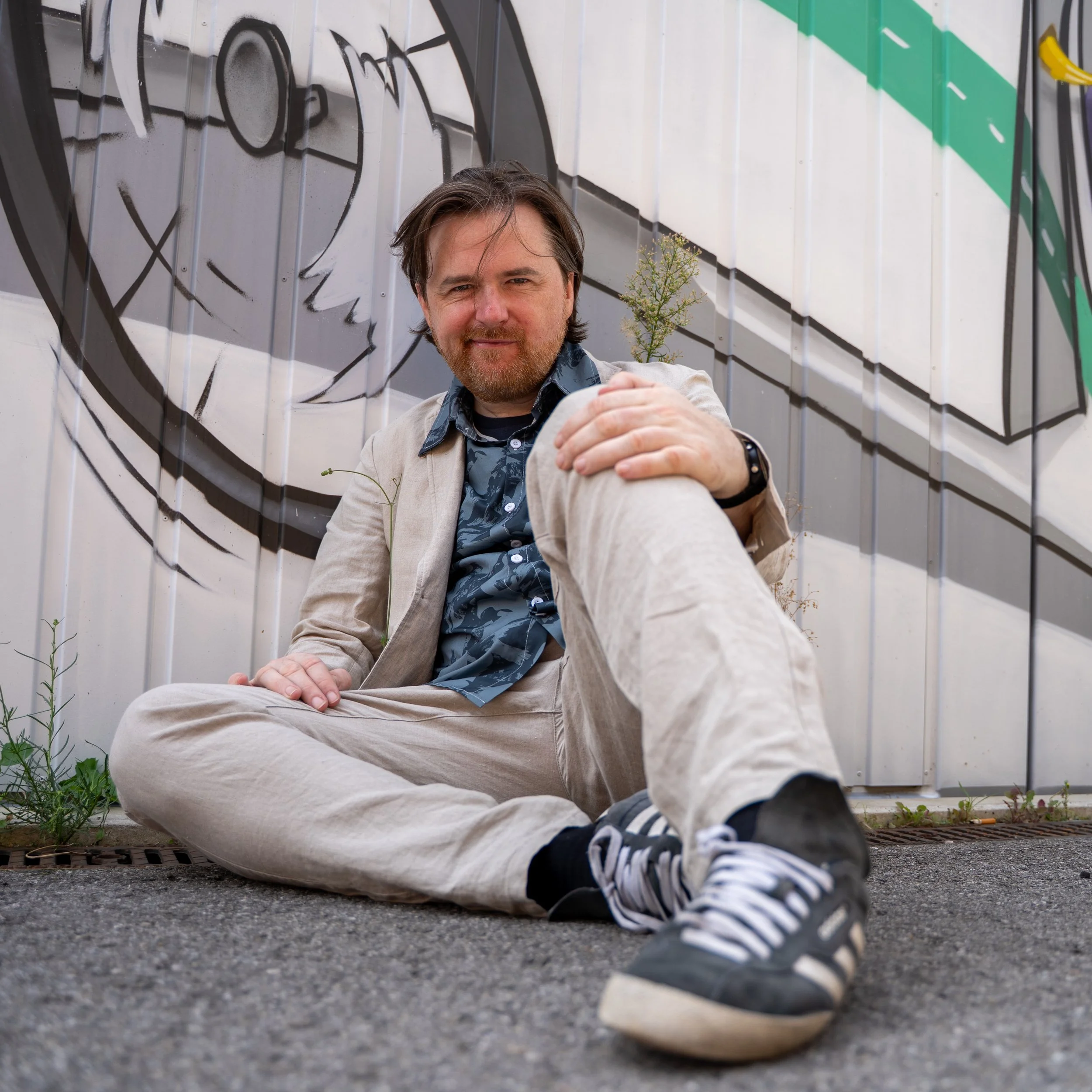 A man with brown hair and a beard sitting on the ground in front of a wall with graffiti art, wearing a beige suit, a blue patterned shirt, black shoes with white laces, and a watch, smiling at the camera.
