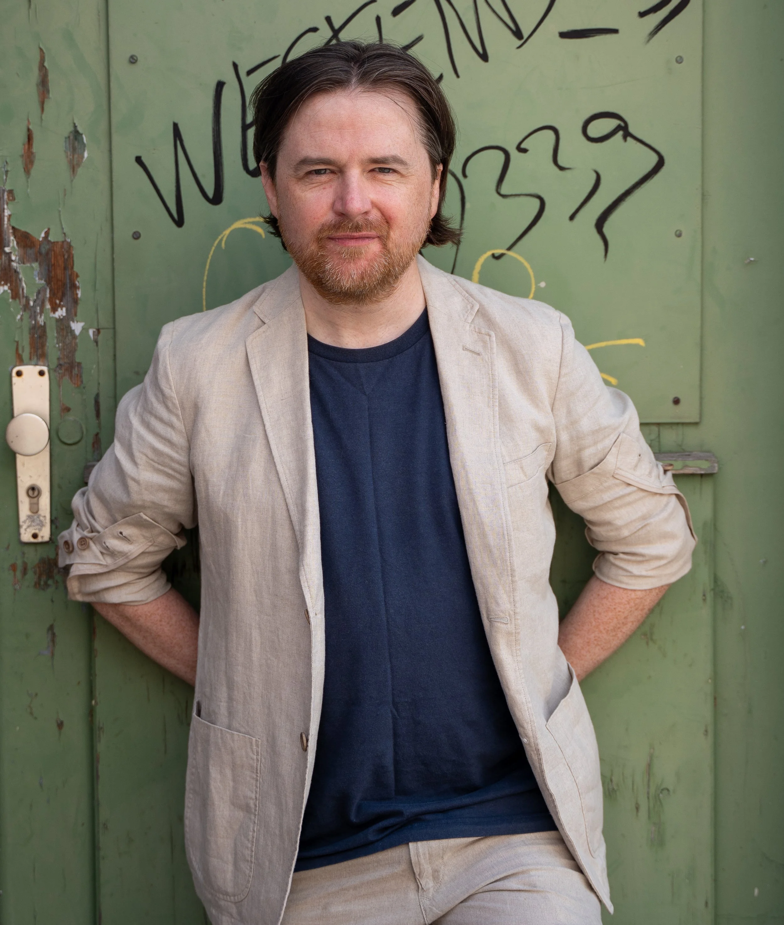 A man with brown hair, a beard, and fair skin, wearing a beige blazer and navy shirt, stands with his hands in his pockets in front of a green wall with graffiti.