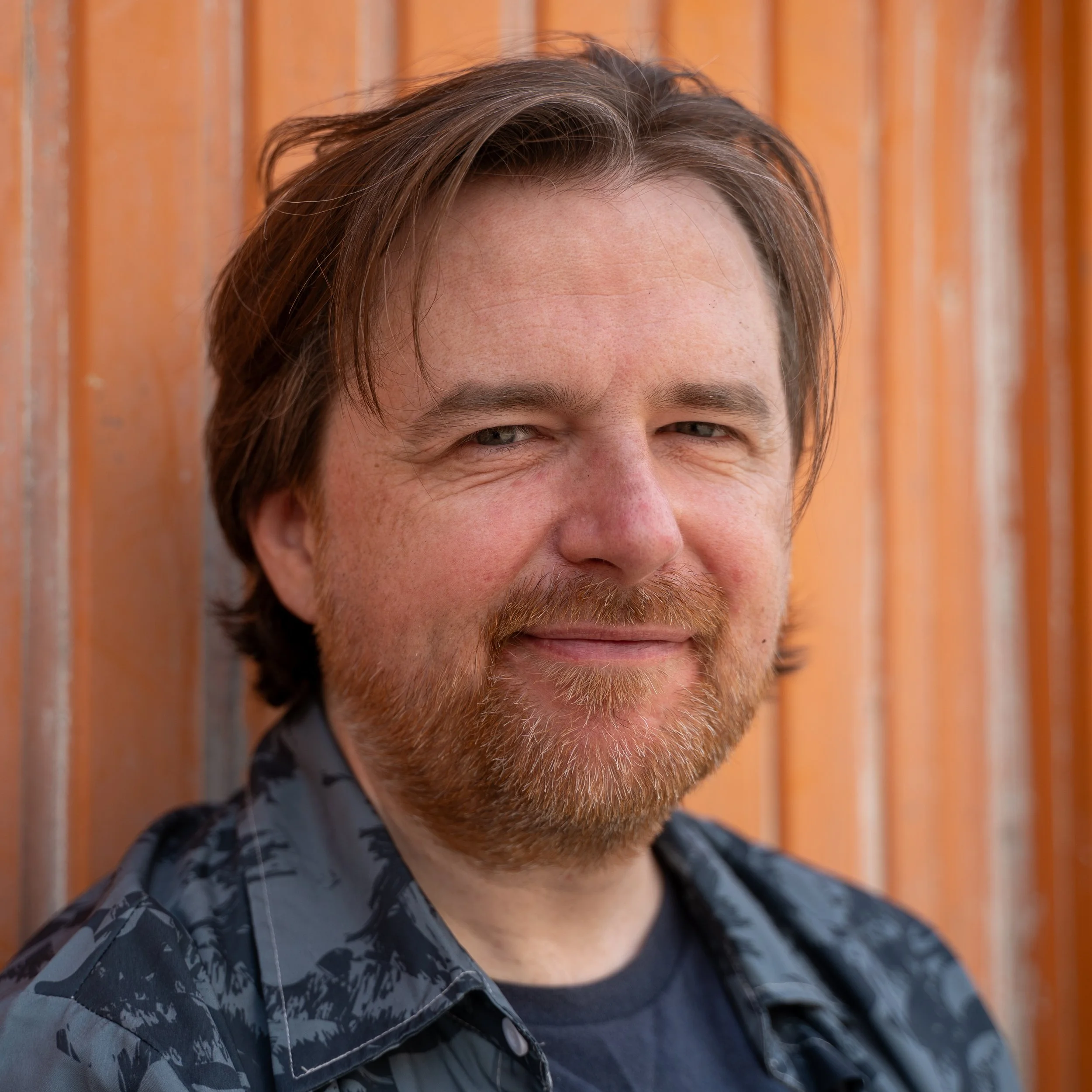 Close-up of a man's face with brown hair, beard, and eyes, smiling slightly, in front of a wooden background. Daniel Beaver