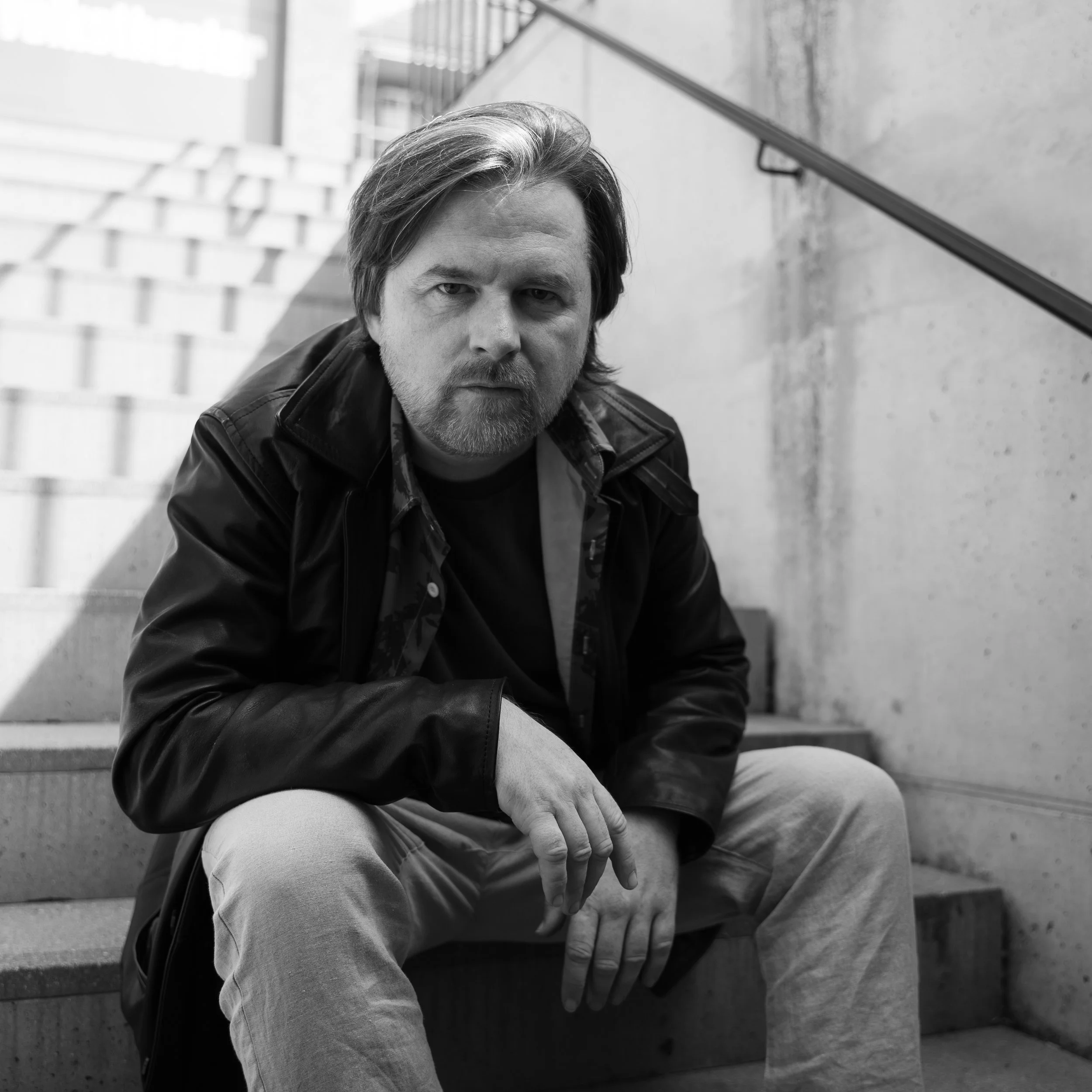 A black and white photo of a man with medium-length hair, a beard, and mustache, sitting on outdoor stairs. He is wearing a leather jacket over a shirt and light-colored pants, looking directly at the camera with a serious expression.