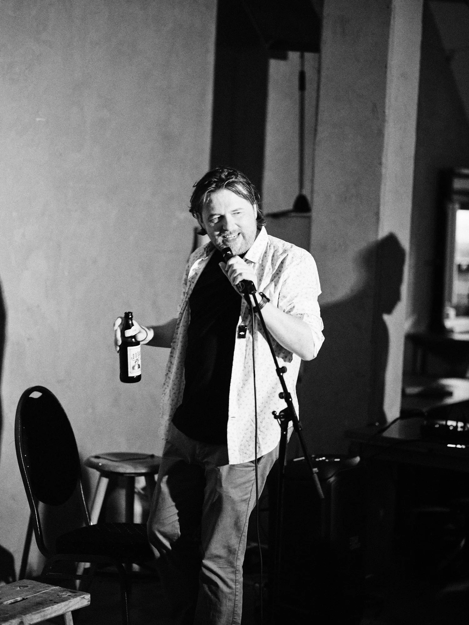 A man with dark hair, a beard, and a mustache holding a bottle in his right hand, standing near a microphone, smiling, in an indoor setting with chairs, a window, and a lamp in the background.