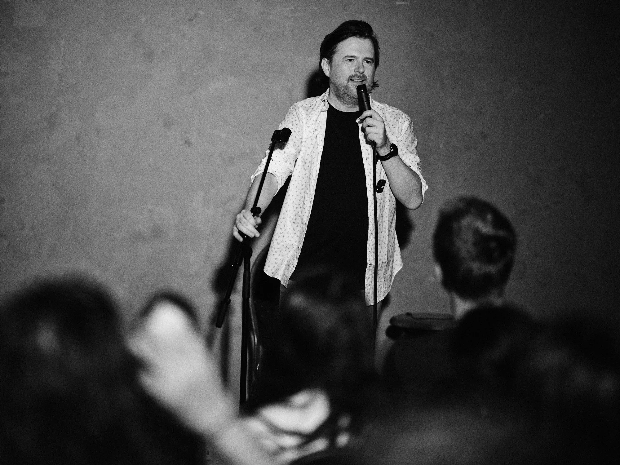 A man with dark hair and a beard, holding a microphone, standing on a stage with a plain background, possibly giving a speech or performance.