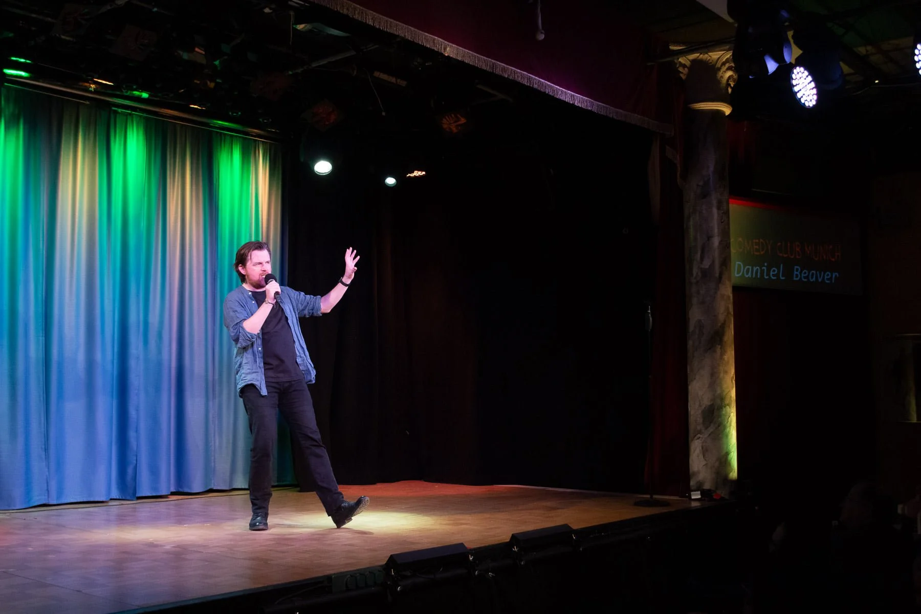 A man performing stand-up comedy on stage with a colorful curtain backdrop. He holds a microphone and gestures with one hand, standing with one foot slightly raised. The stage has a wooden floor and lighting from above.