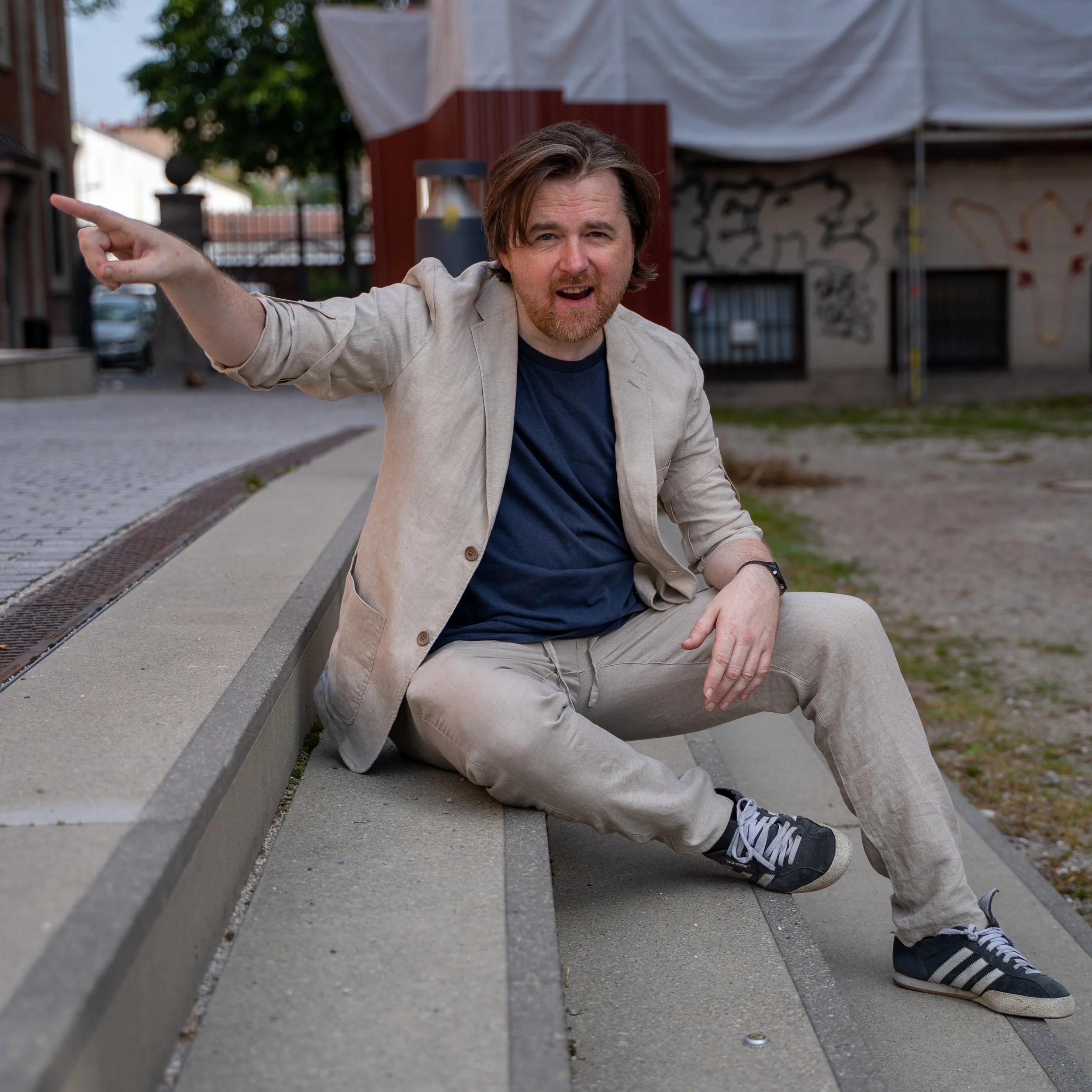 A man with brown hair and a beard sitting on a concrete curb, pointing to the left and smiling, wearing a beige blazer, navy blue t-shirt, beige pants, and black sneakers with white stripes, in an urban setting with graffiti on the wall behind him.