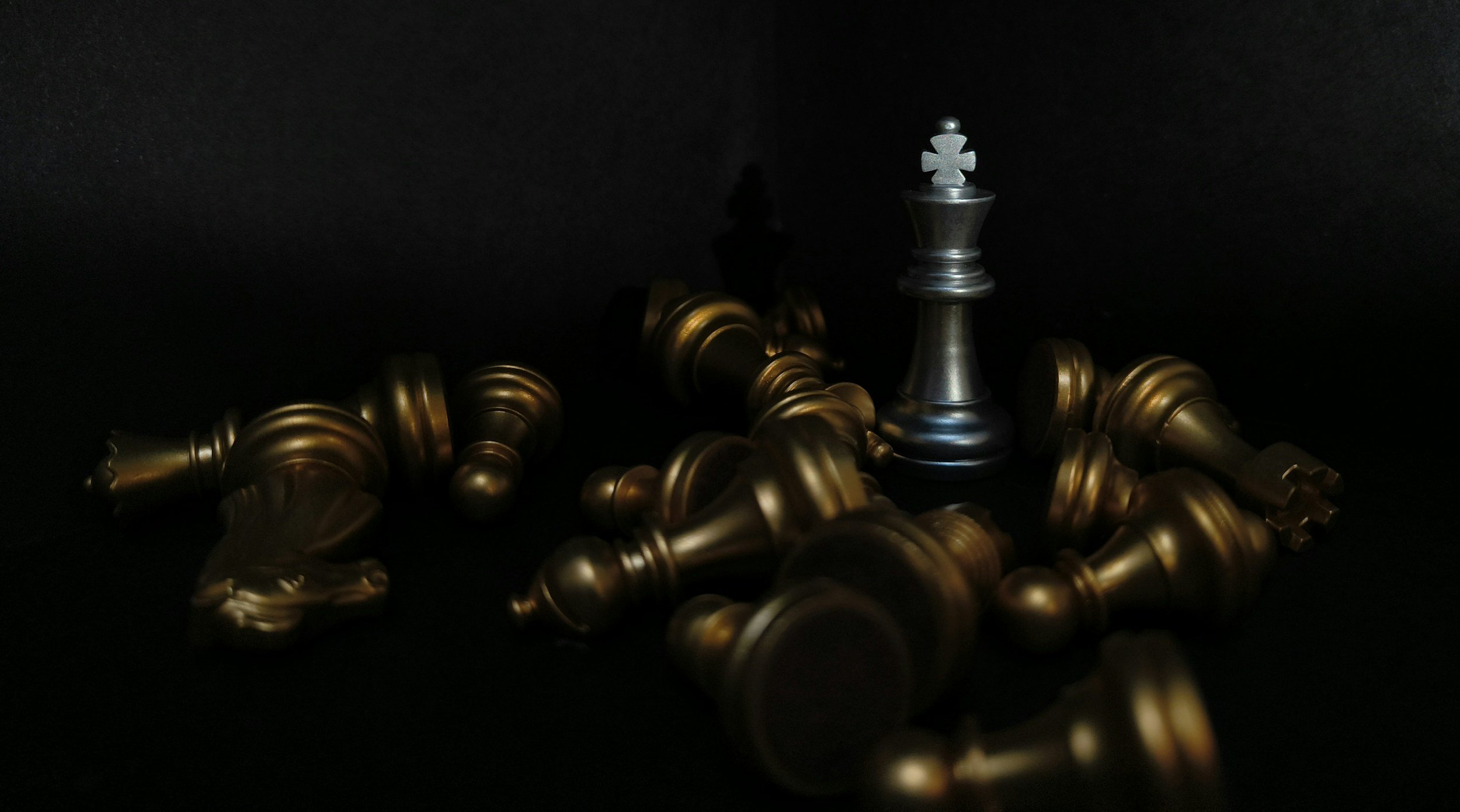 A silver king chess piece standing upright among fallen gold chess pieces on a dark surface.