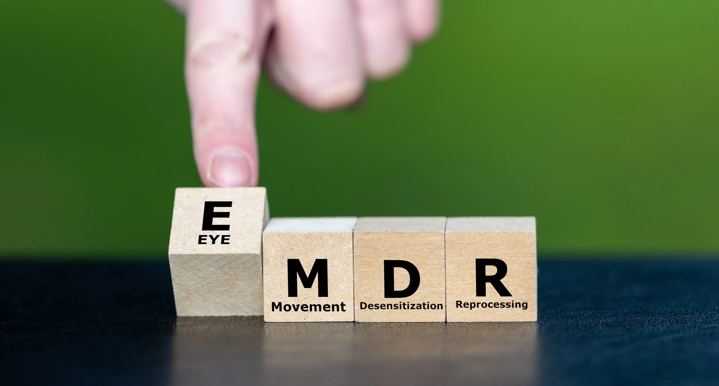 Wooden blocks spelling out 'EYE', 'M' for Movement, 'D' for Desensitization, and 'R' for Reprocessing, with a person's finger pointing at the 'EYE' block.