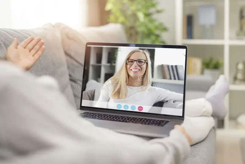 Person lying on couch having a video call on a laptop with a smiling woman on the screen.