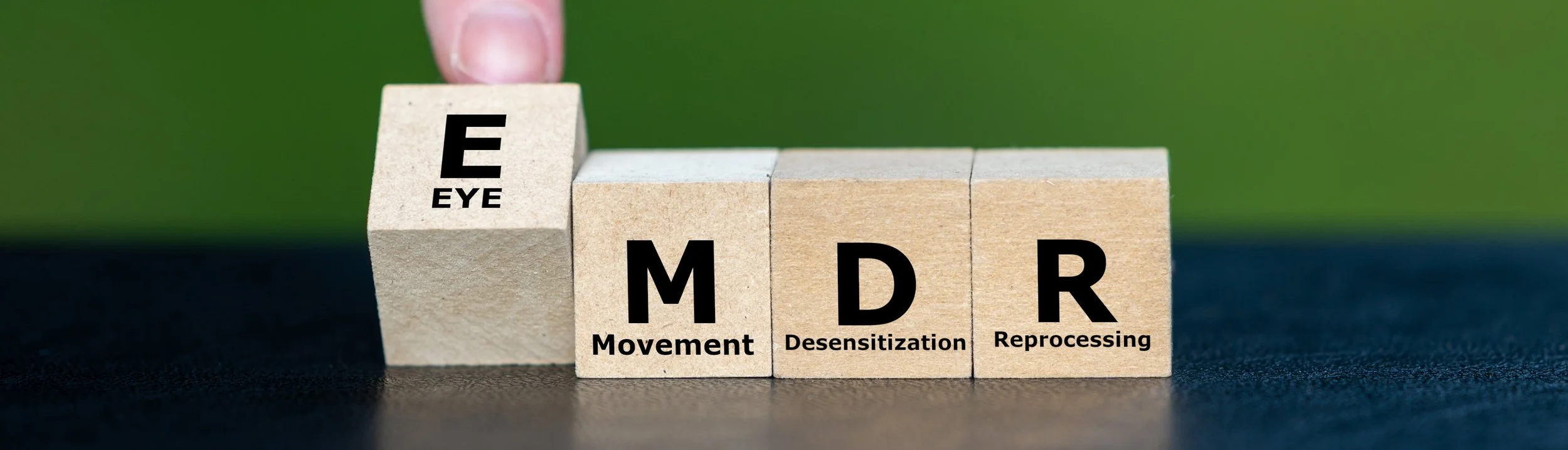 Wooden blocks spelling out E M D R with words 'Eye', 'Movement', 'Desensitization', and 'Reprocessing' printed on them, arranged in a row on a dark surface with a green background.