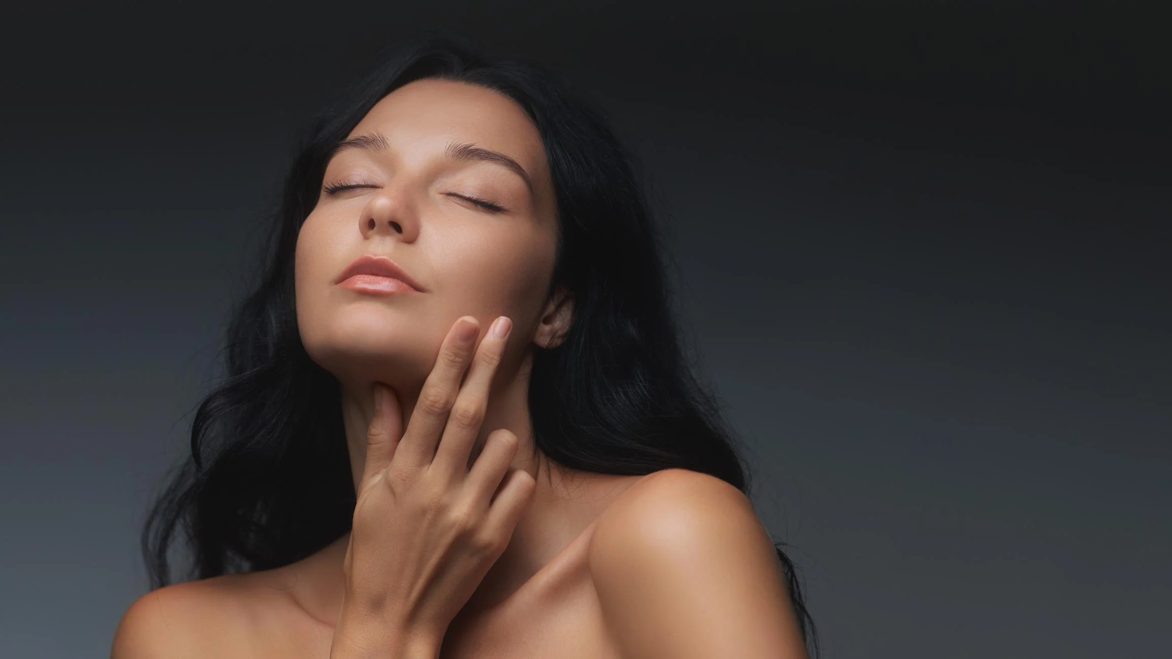 A woman with long dark hair and light skin has her eyes closed, touching her face gently with her hand, against a dark gray background.
