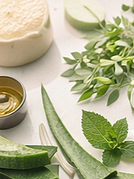 Aloe vera leaf, mint leaves, cucumber slices, a small bowl of oil, and a jar of cream on a white surface.