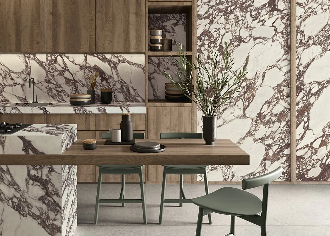 A modern kitchen featuring Lea Ceramiche's Noblesse Viola Arabesque porcelain surfaces, complemented by warm wood cabinetry and matte green chairs, available through PietraCasa.