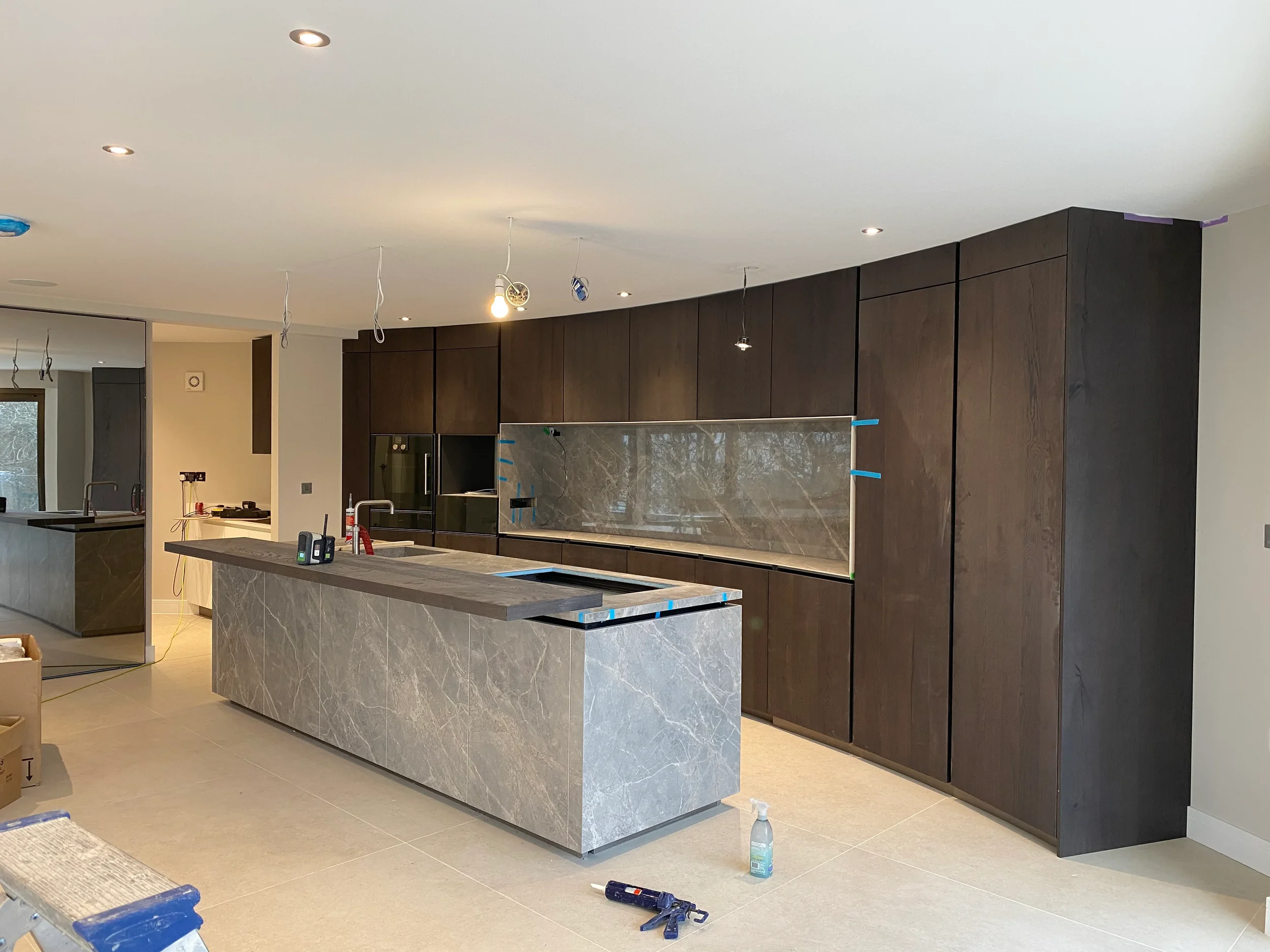 Contemporary kitchen installation under construction featuring a large monolithic island and matching full-height splashback niche custom fabricated from grey veined marble-effect porcelain slabs, paired with floor-to-ceiling dark wood cabinetry.