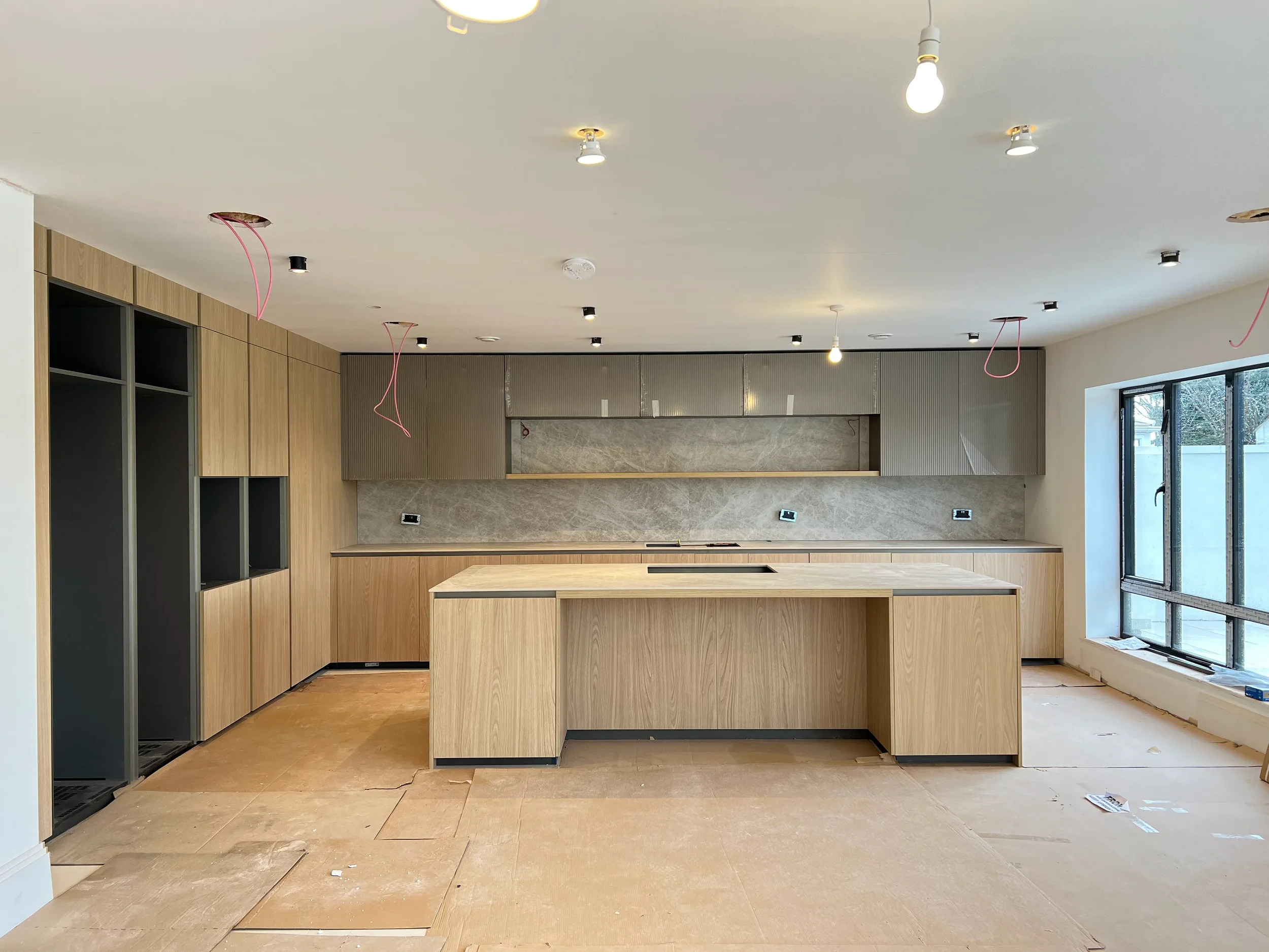 High-end contemporary kitchen site highlighting custom architectural porcelain fabrication, showcasing seamless porcelain slab wall cladding behind minimalist oak cabinetry and an expansive central island display with temporary protective flooring.