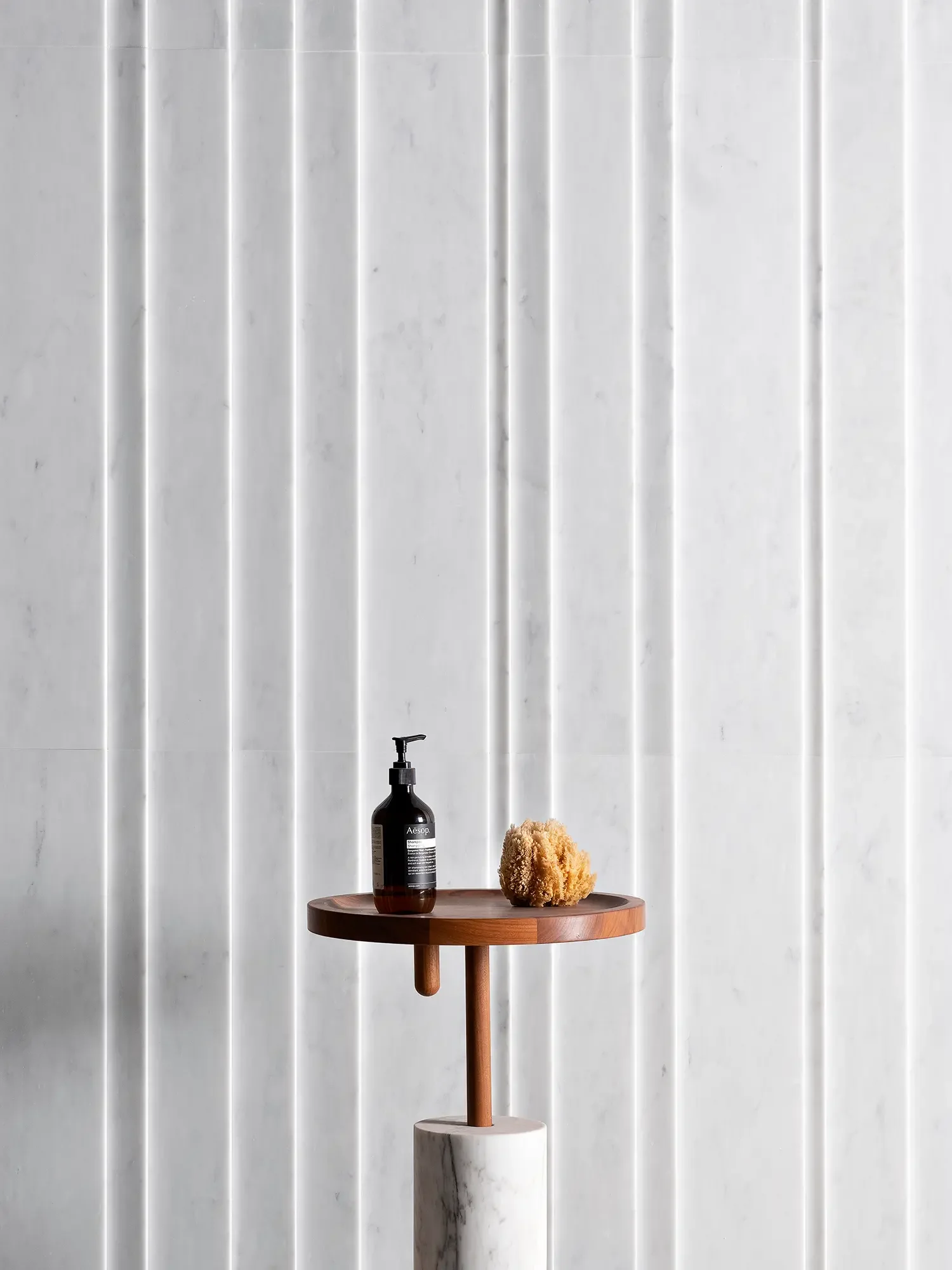 The Luce di Carrara Underground column, a luxury bathroom or vanity accessory crafted from solid marble and wood, featuring a round, rotating tray on a cylindrical marble base, available through PietraCasa.