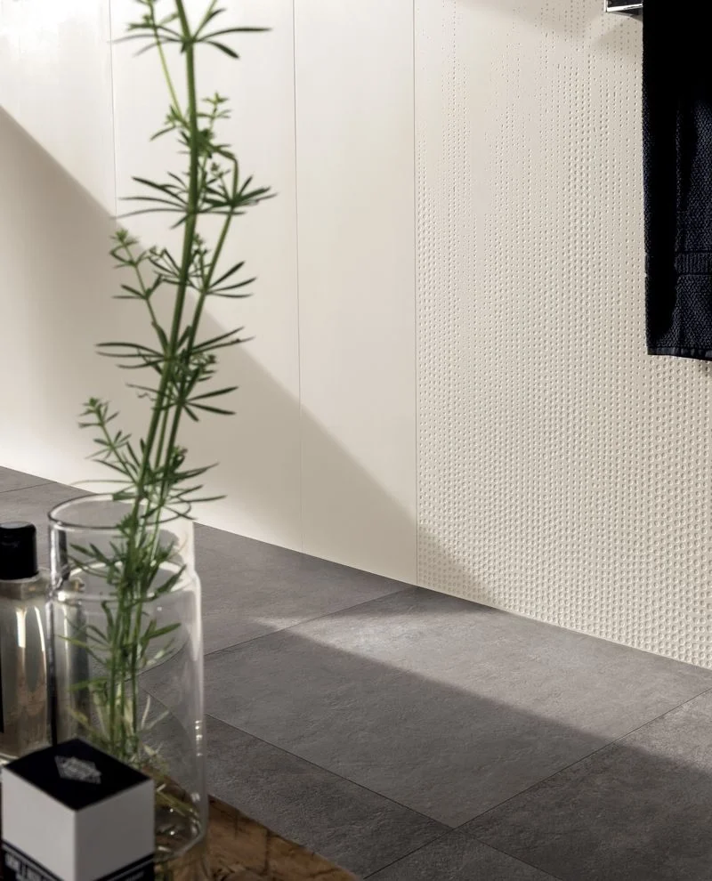 A minimalist bathroom interior featuring the Talco collection by Kronos Ceramiche, showcasing a feature wall with plain and textured 3D decorative tiles, such as 'Dégradé' or 'Lines,' paired with coordinating dark, concrete-look porcelain floor tiles