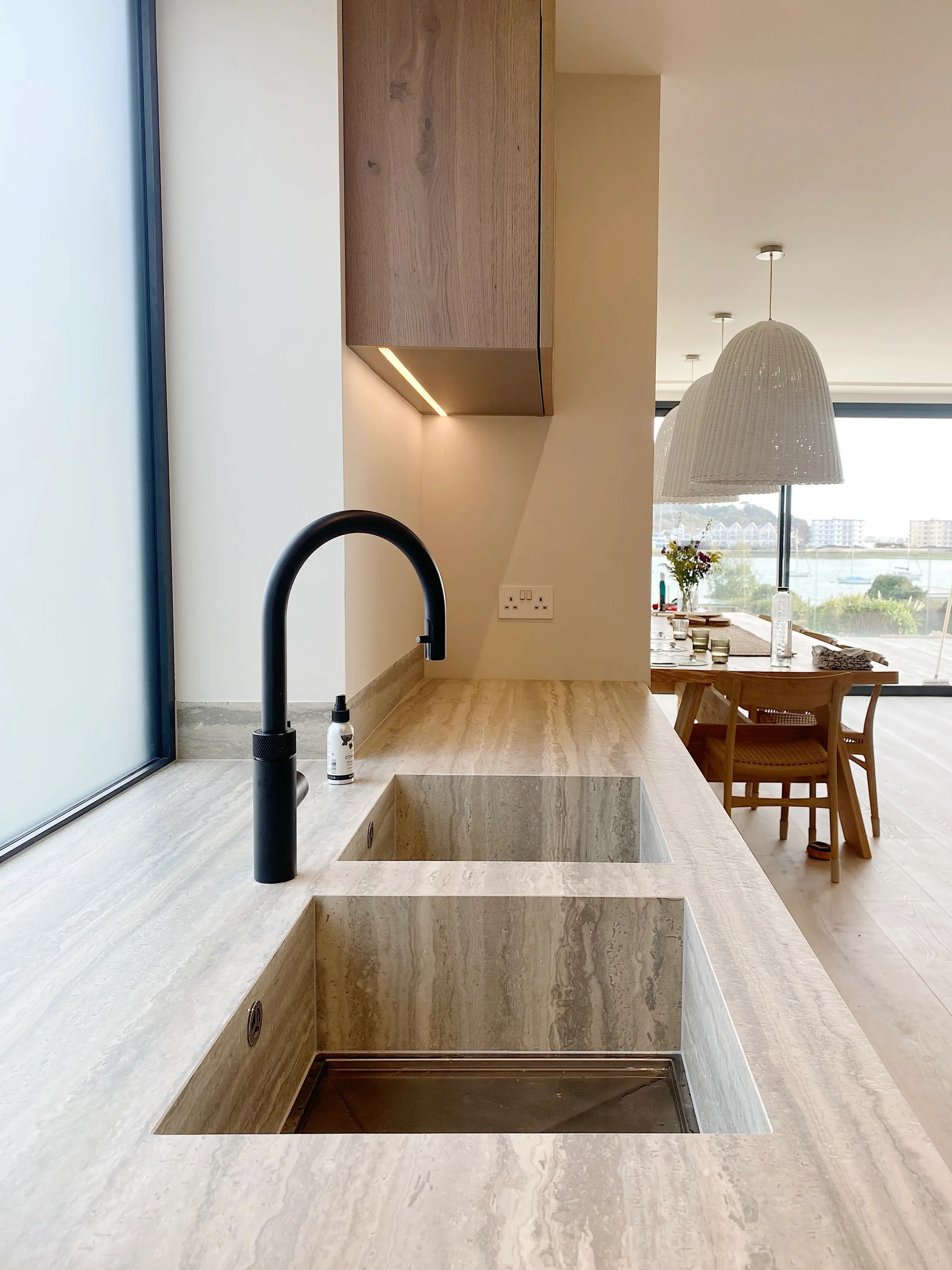 Luxury kitchen installation featuring a bespoke double integrated sink and matching worktop custom fabricated from vein-cut travertine-effect porcelain slabs, paired with a contemporary matte black swan-neck tap and minimalist oak cabinetry.