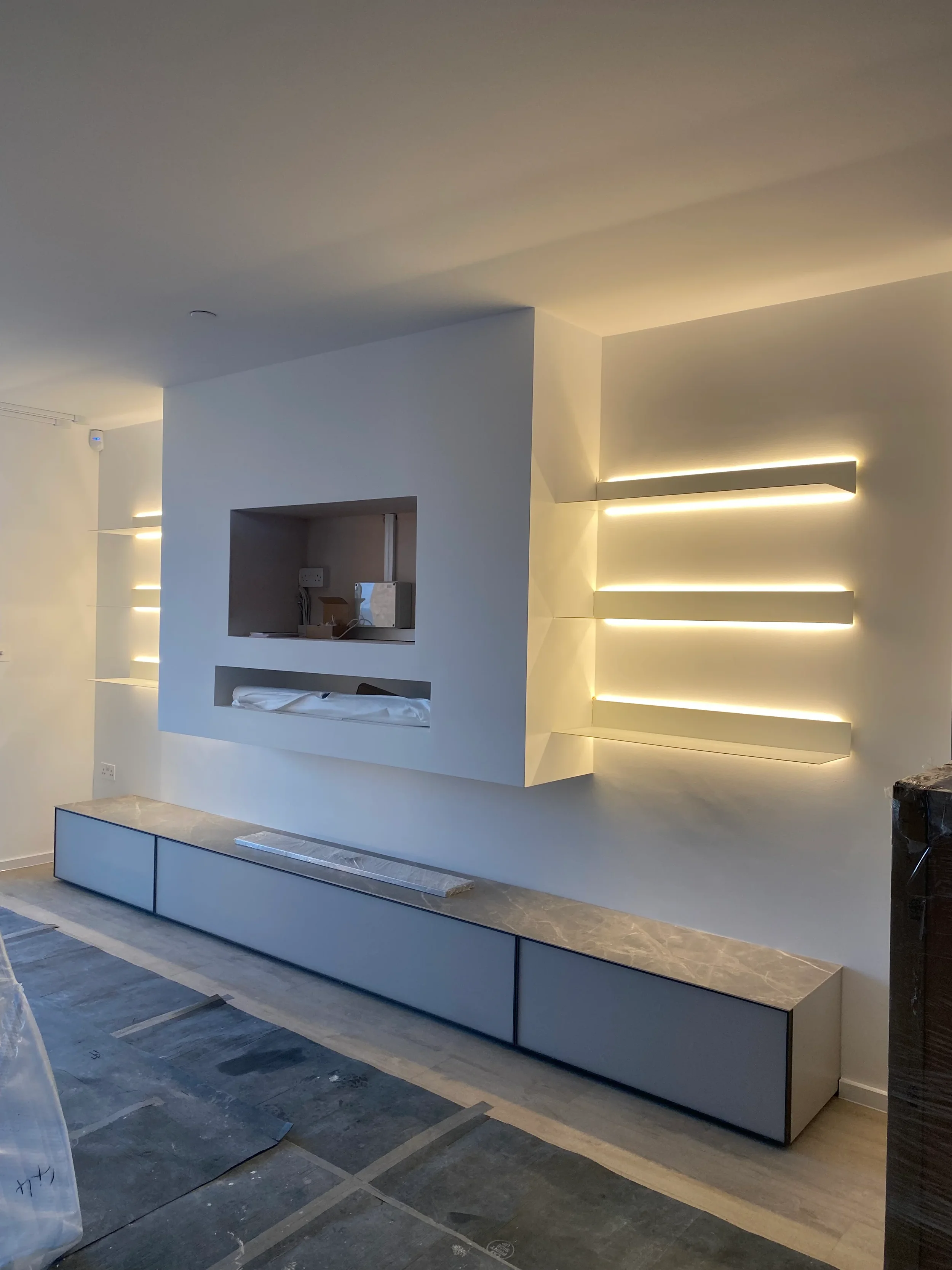 High-end minimalist living room design under construction, featuring a floating chimney breast alcove for a TV and fireplace, flanked by custom floating shelves with integrated LED strip lighting.