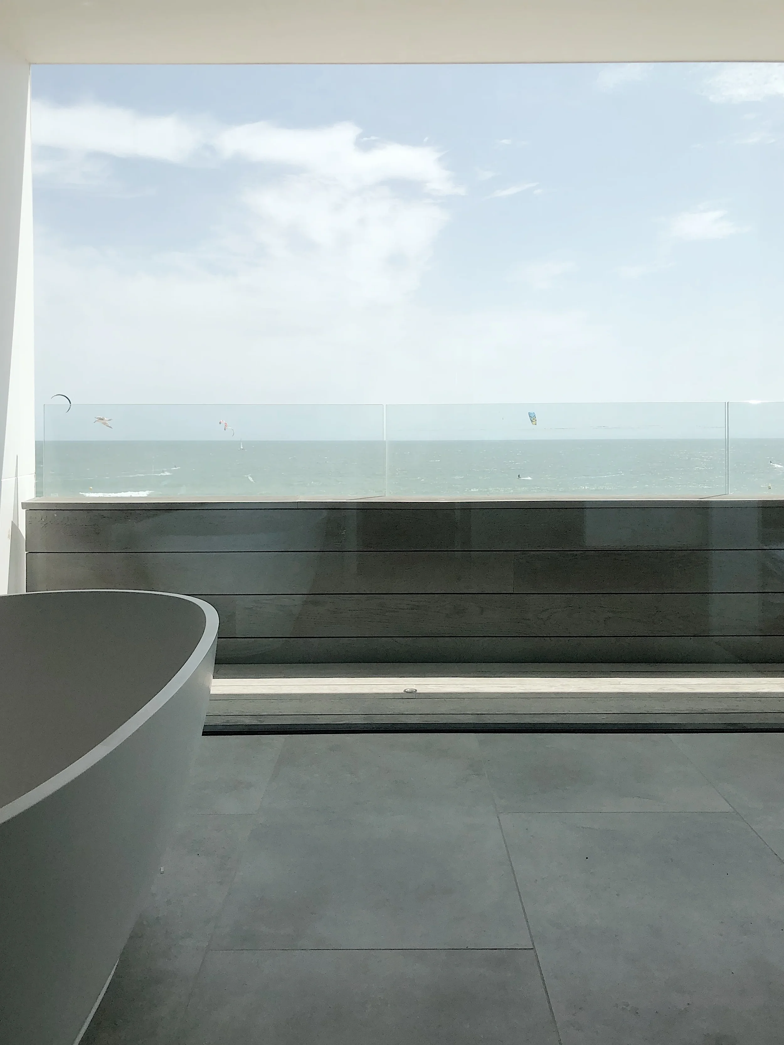 Modern open-plan bathroom with a freestanding tub, grey porcelain flooring, and a glass balcony balustrade with sea views