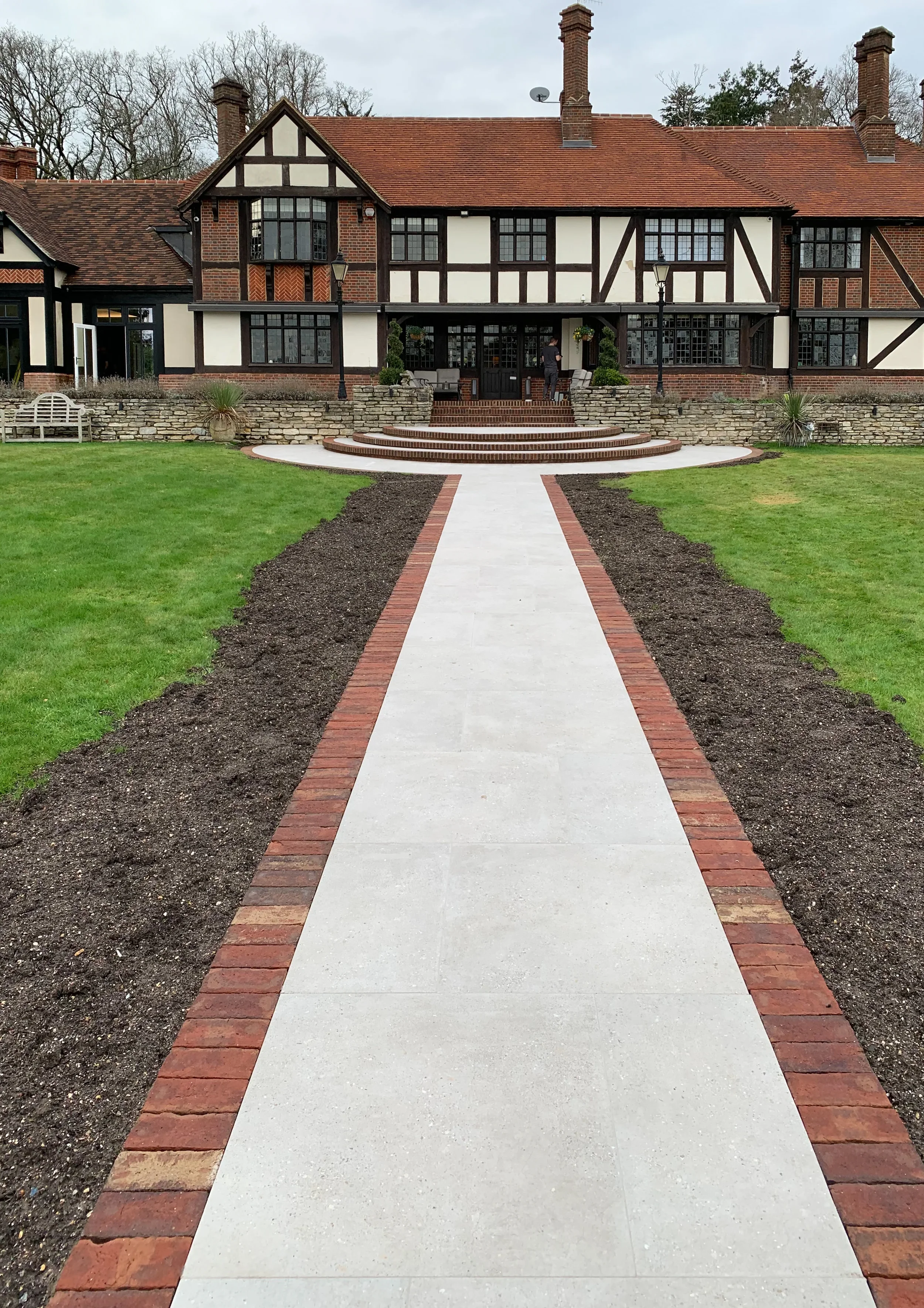 Garden walkway featuring light grey porcelain paving stones framed by traditional red brick edging, set within a landscaped lawn area.