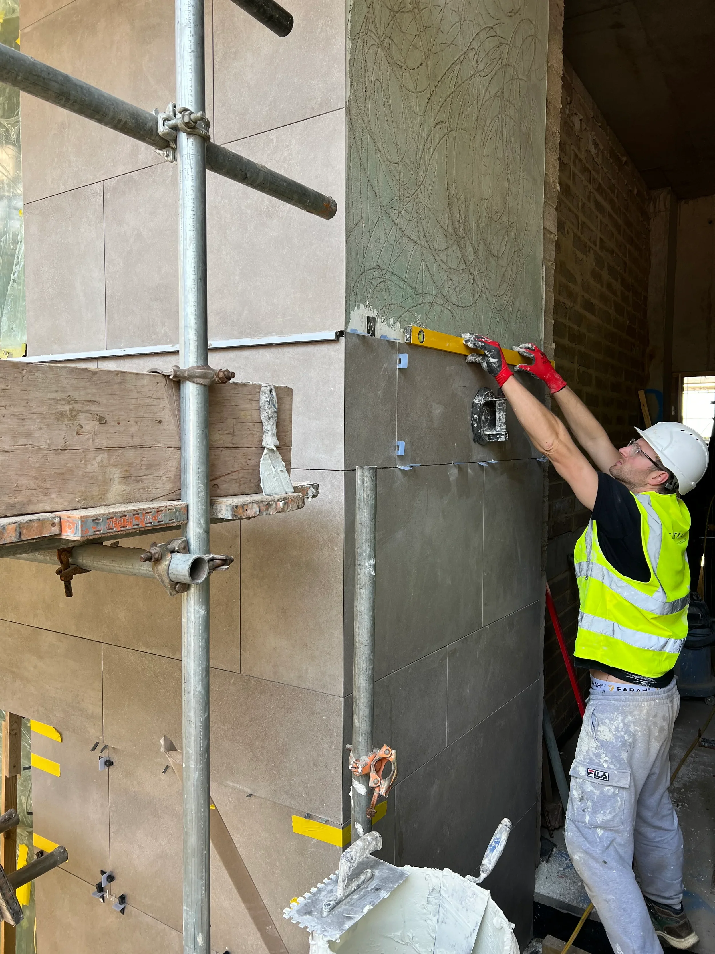 Professional tiler installing Keope Moov Grey large-format porcelain slabs on an architectural feature wall or column using a spirit level, 1mm levelling clips, and Peygran tile levelling wedges.