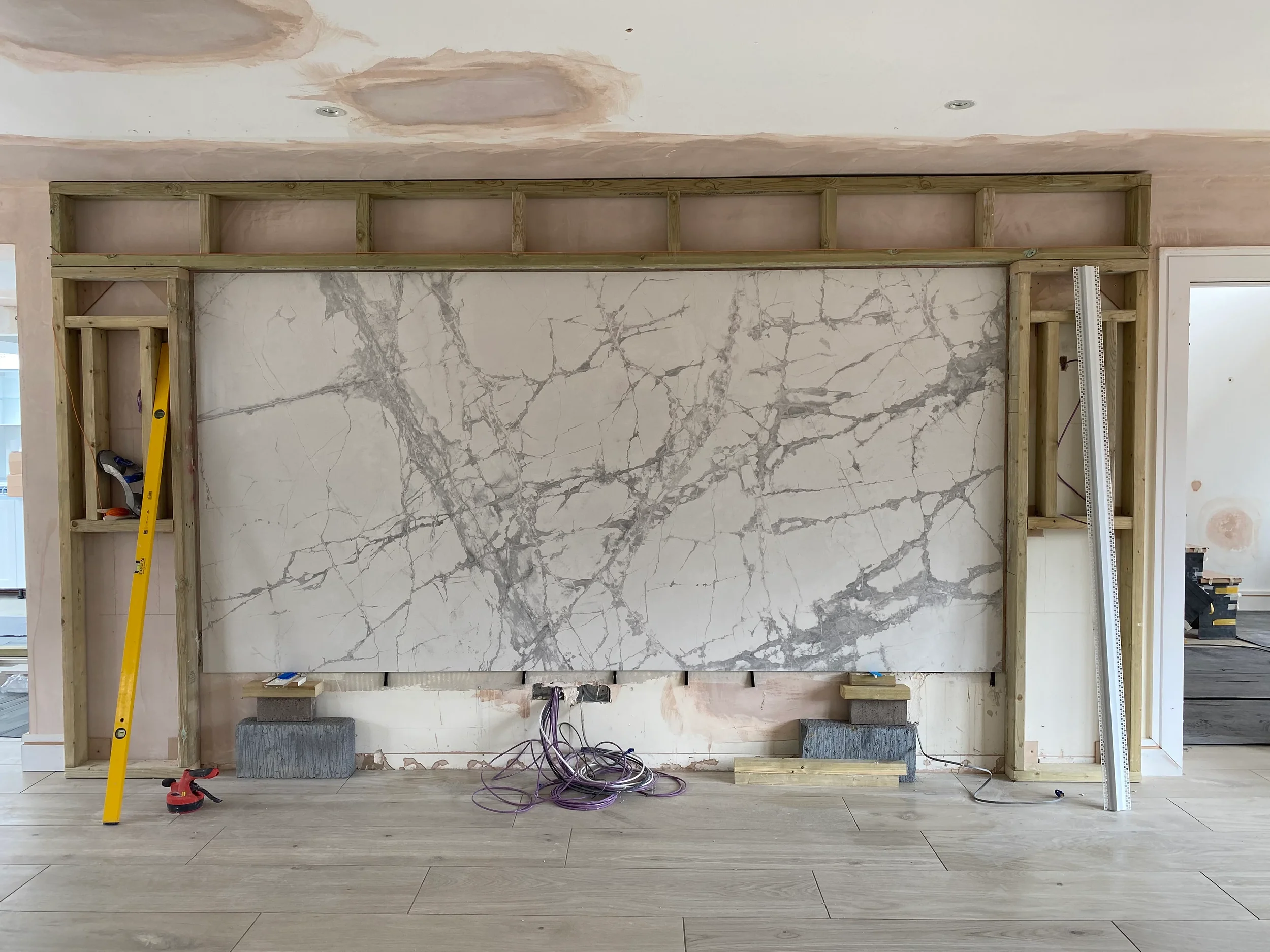 An interior renovation project showcasing the structural timber framework of a custom media wall, featuring a large-format marble-effect porcelain panel installation in progress.