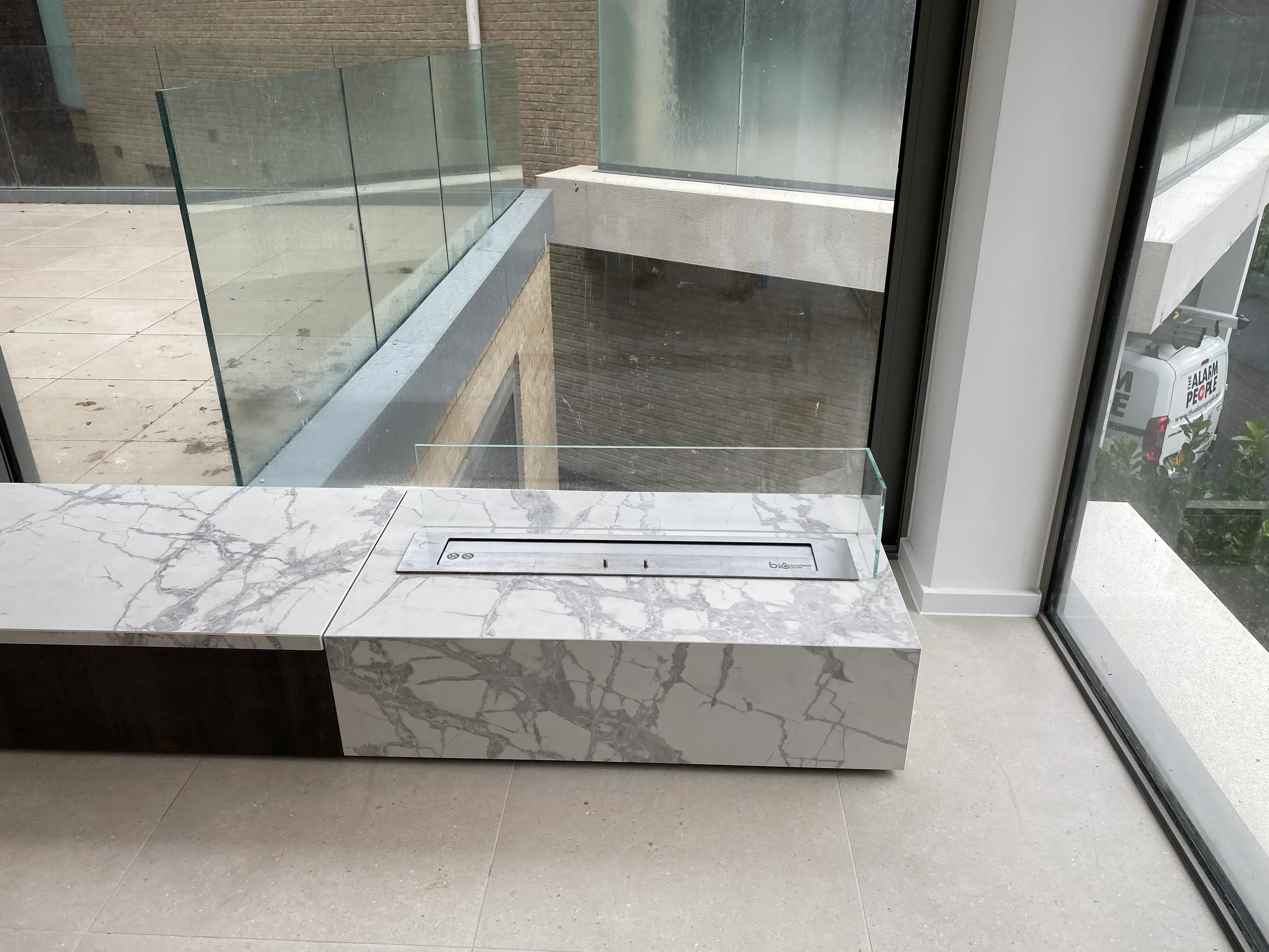 A luxury contemporary low-profile bench custom-fabricated from heavily veined white marble-effect porcelain slabs, featuring an integrated linear bioethanol burner with a protective glass screen.